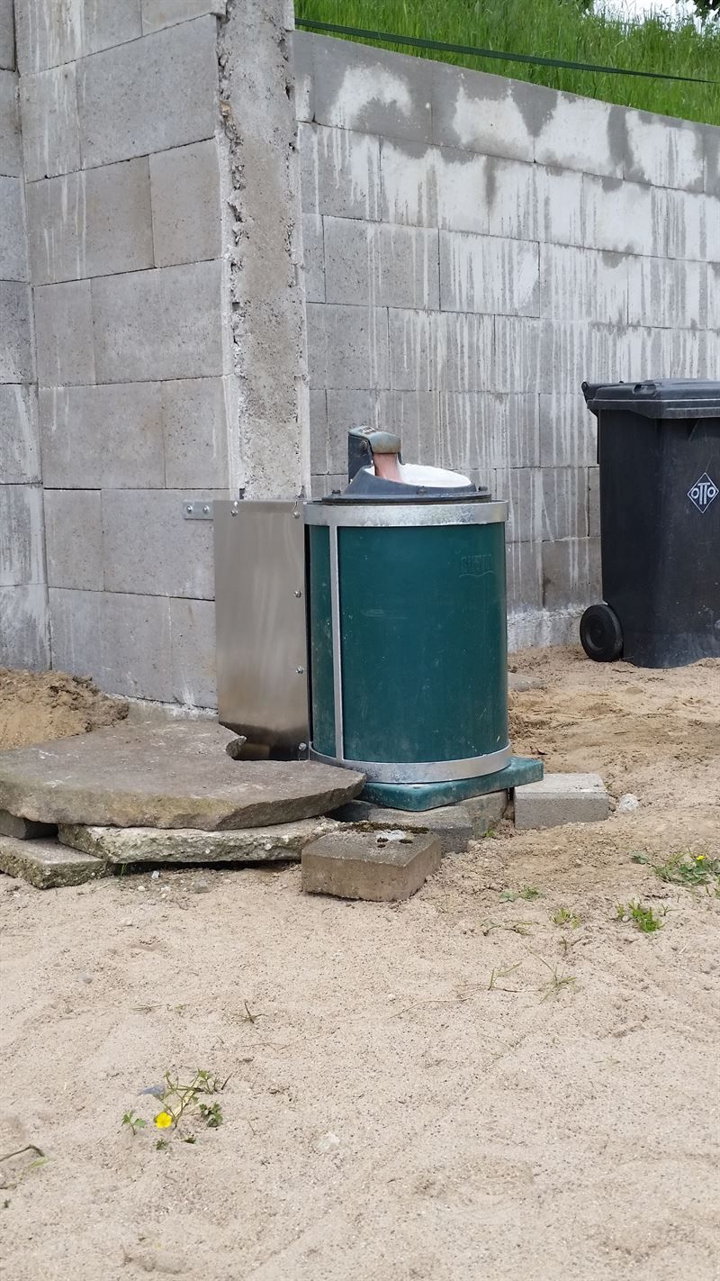 Eine grüne Mülltonne steht im Dreck vor einer Backsteinmauer.