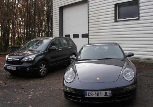 Deux voitures garées devant un bâtiment, un SUV noir et une Porsche grise devant un garage.