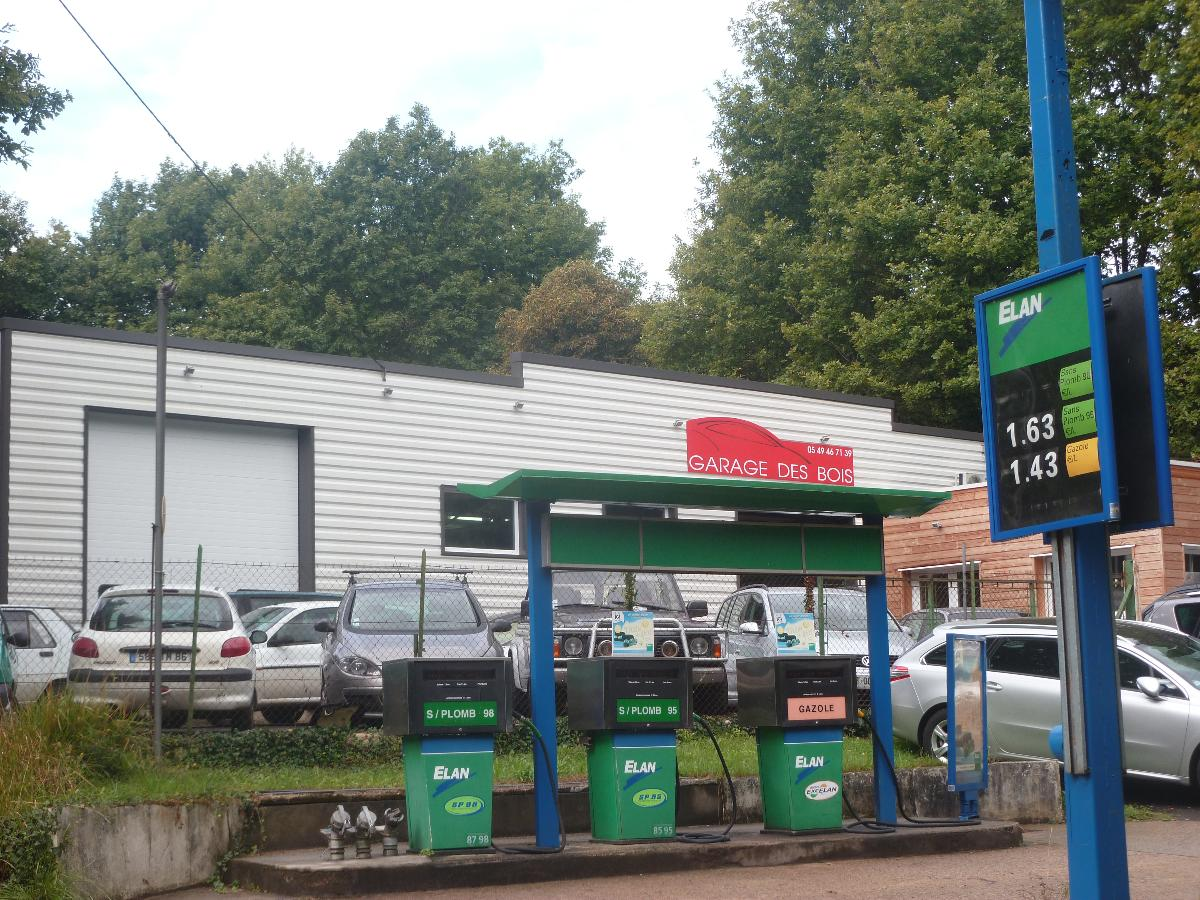 Station-service avec des pompes sous un auvent vert et un panneau de prix, à côté d’une voiture blanche garée