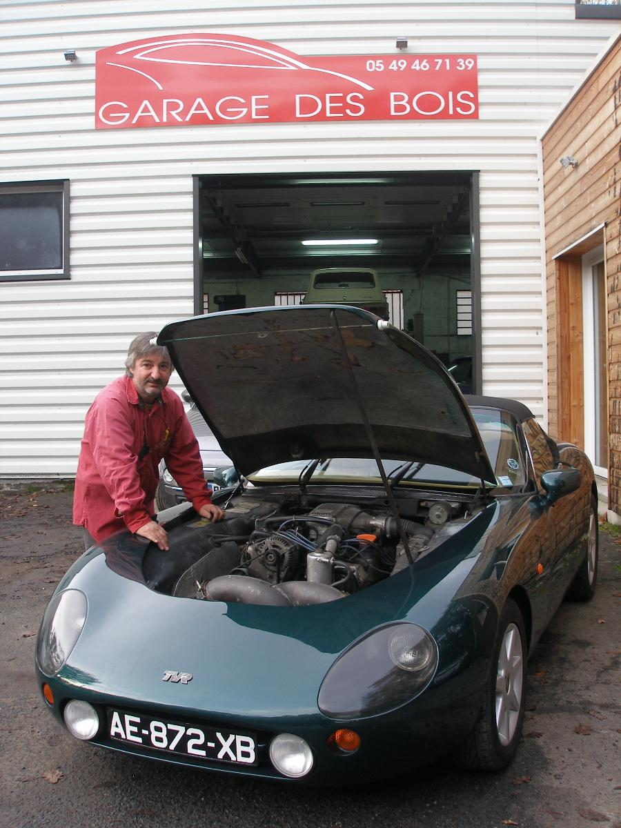Un mécanicien à côté d’une voiture de sport vert foncé au capot ouvert, devant Garage des Bois