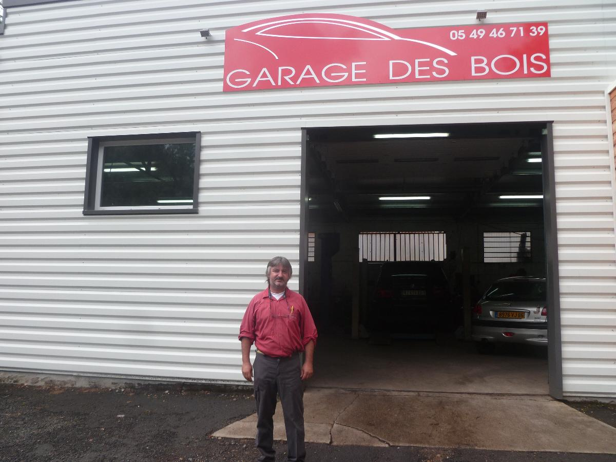 Une personne se tient devant un garage, sous une enseigne rouge « GARAGE DES BOIS » au-dessus de l’entrée