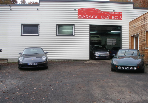 Deux voitures de sport noires garées devant un garage avec une enseigne rouge où l’on peut lire « Garage des Bois »