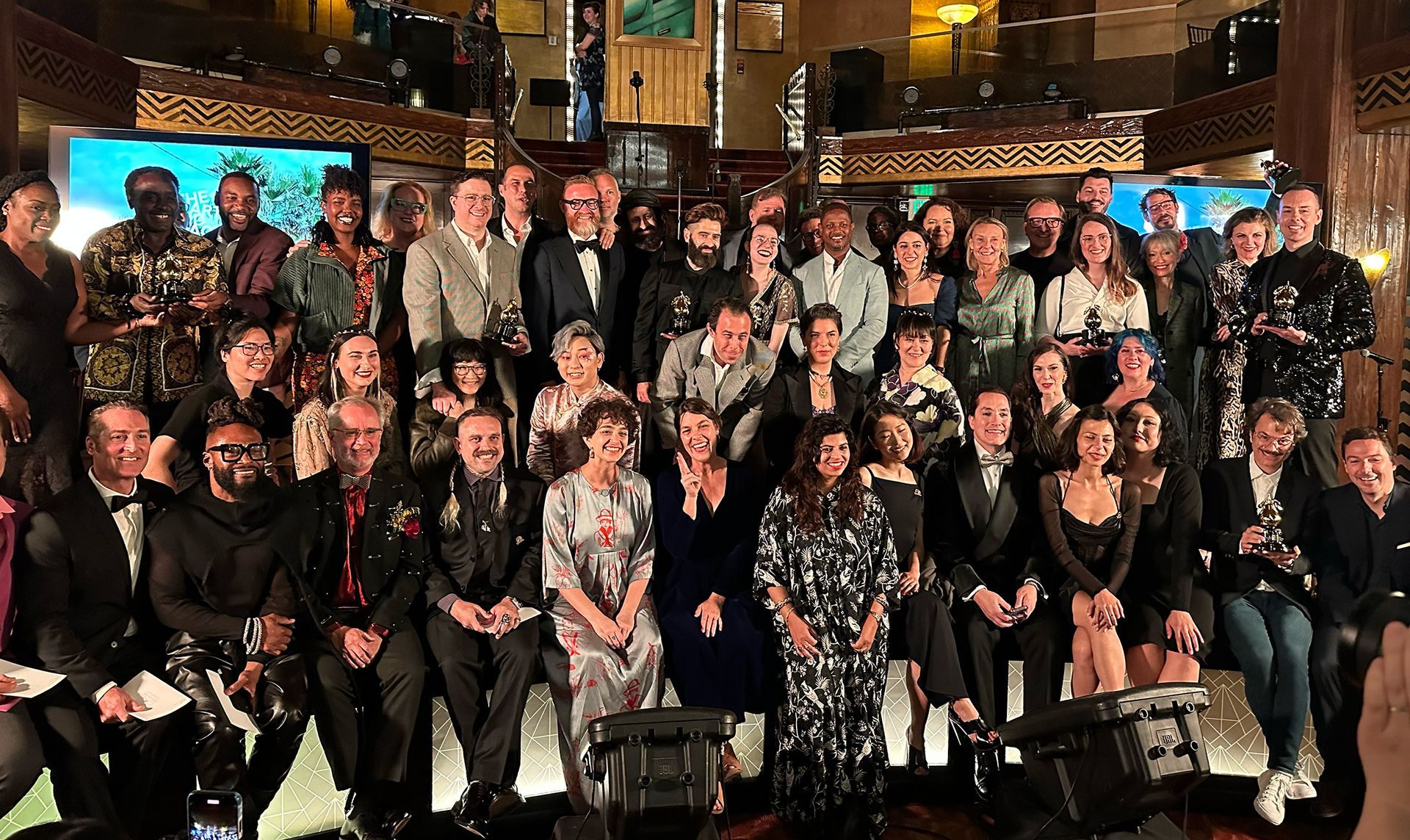 Un numeroso grupo de personas posa para un evento formal en un salón de baile adornado con oro, bajo una lámpara de araña de cristal.