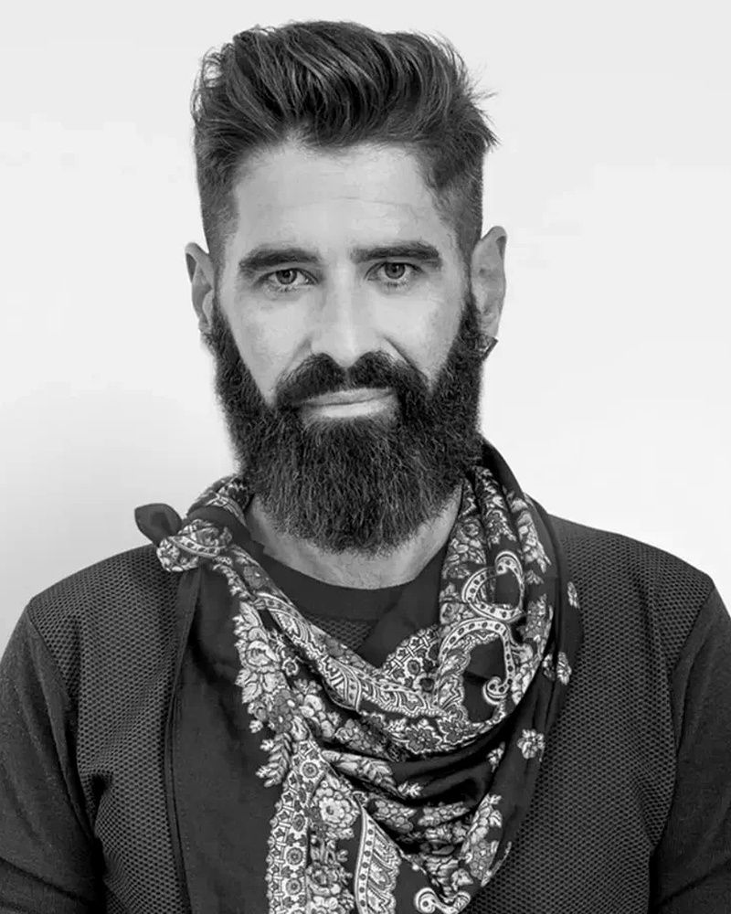 Hombre con barba poblada y bufanda estampada, mirando al espectador.