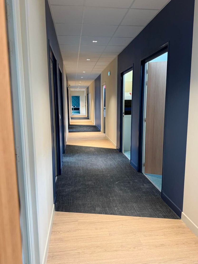 Couloir étroit de bureaux aux murs bleus, au sol moquetté et bordé de plusieurs portes de chaque côté