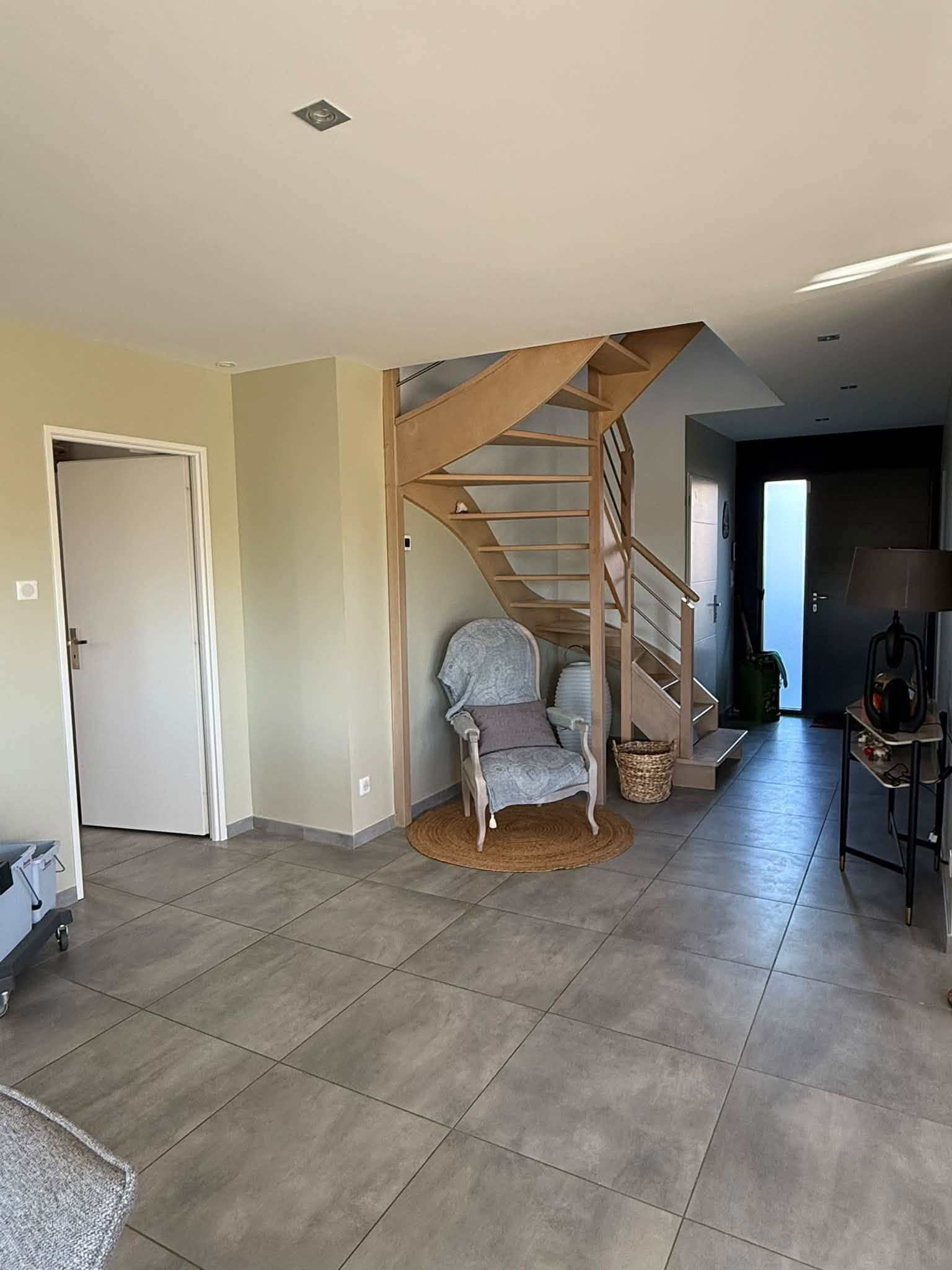 Couloir intérieur avec un sol carrelé gris, un escalier en bois et une petite chaise sous l’escalier