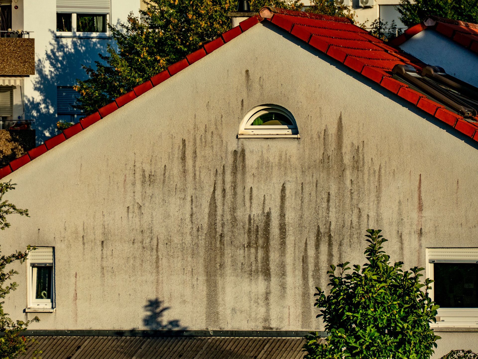 Mur blanc patiné d’une maison au toit rouge, avec une petite fenêtre en arc, en partie ombragé par des arbres