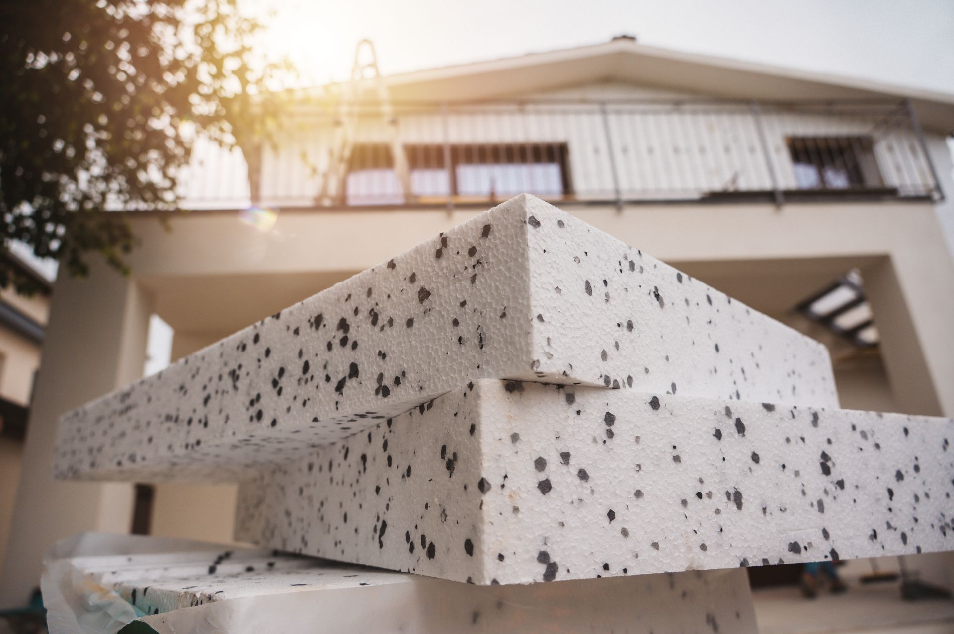 Des blocs de polystyrène isolant empilés, devant une maison à la façade blanche avec balcon