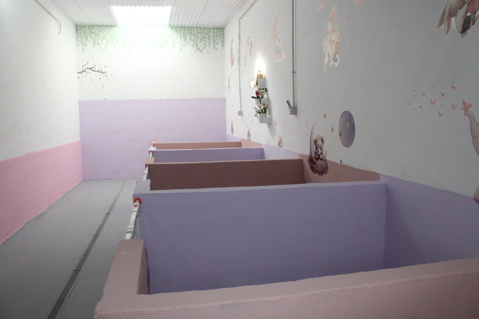 Una habitación vacía con cubículos color lavanda, paredes rosas y blancas, y una claraboya.