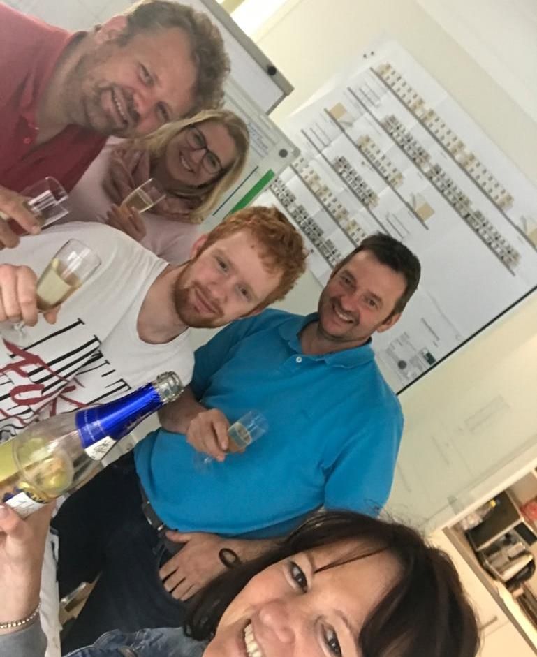 Eine Gruppe von Menschen steht um einen Tisch herum, eine Frau hält eine Flasche Wein