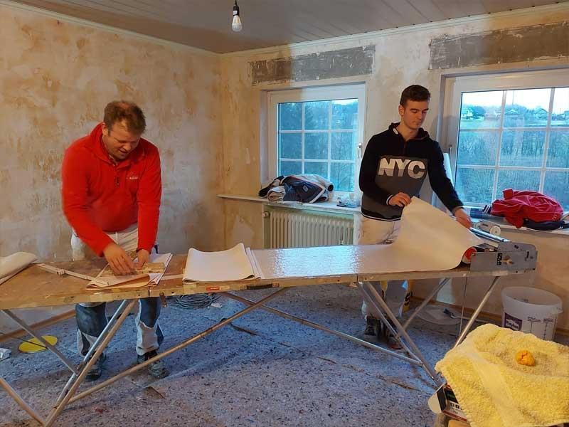 Zwei Männer arbeiten an einem Tisch in einem Raum. Einer der Männer trägt ein NYC-Sweatshirt.