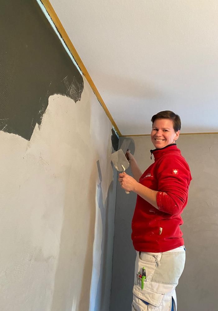 Eine Frau in einer roten Jacke lächelt, während sie eine Wand streicht.