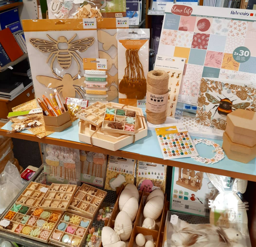 Présentoir de matériel de loisirs créatifs : abeille en bois, papier, ficelle, peintures, objets décoratifs et outils sur des étagères.