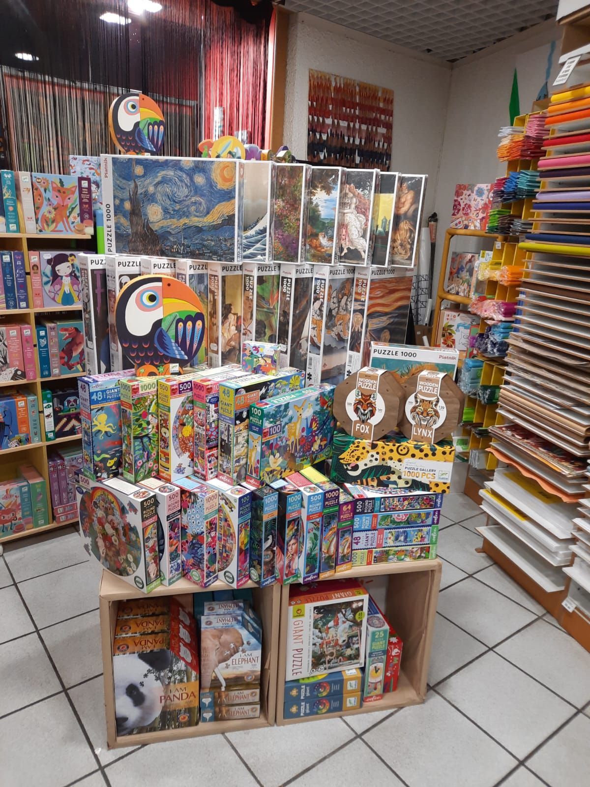 Une vitrine présentant des boîtes de puzzles empilées, dont une grande découpe de toucan, sur des étagères en bois.
