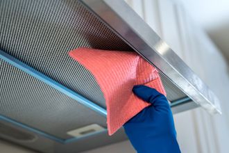 Persona con guante azul limpiando una campana extractora de acero inoxidable con un paño rosa.