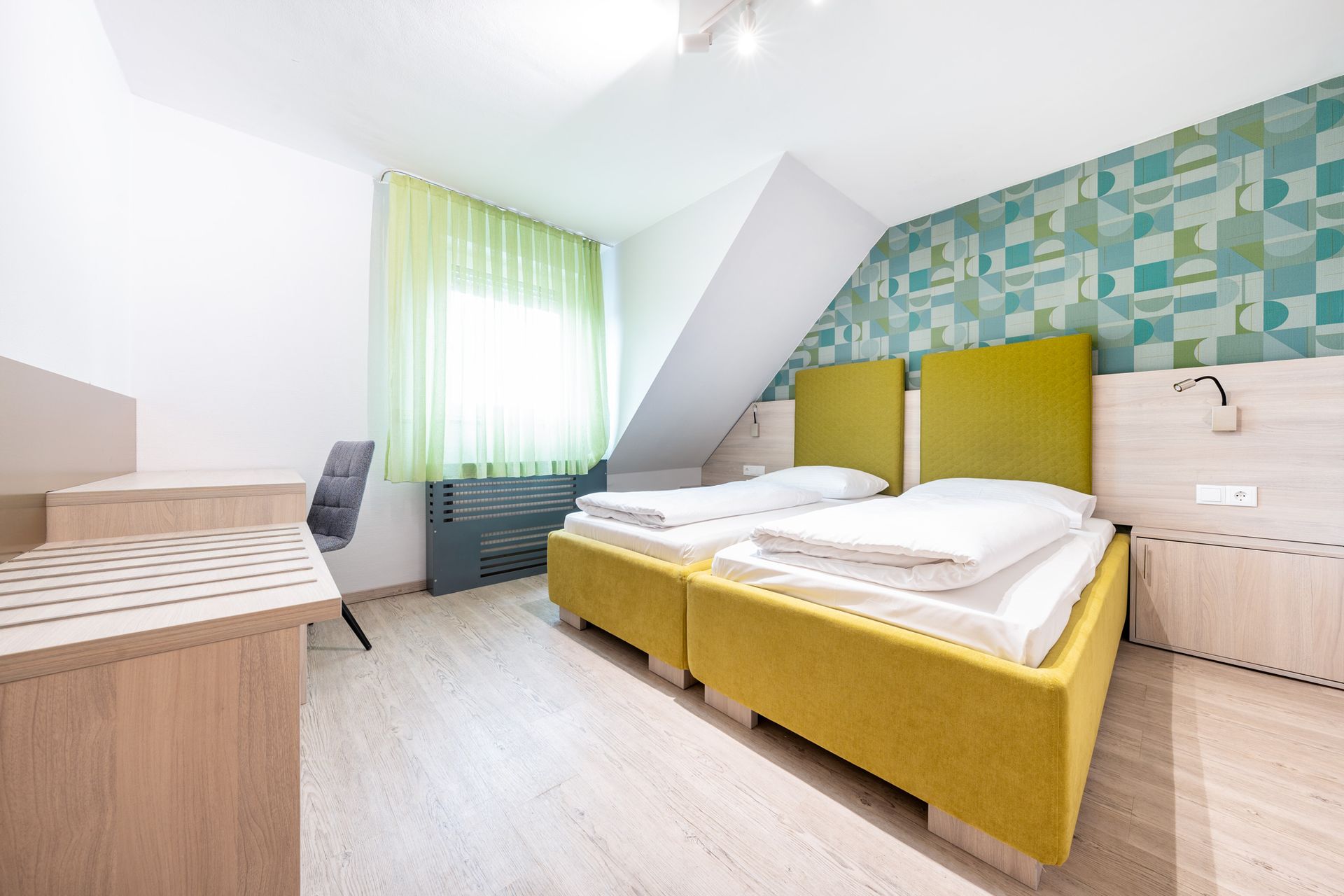 Schlafzimmer mit zwei Betten, gelben Kopfteilen und einer gemusterten Akzentwand. Schreibtisch mit Stuhl.