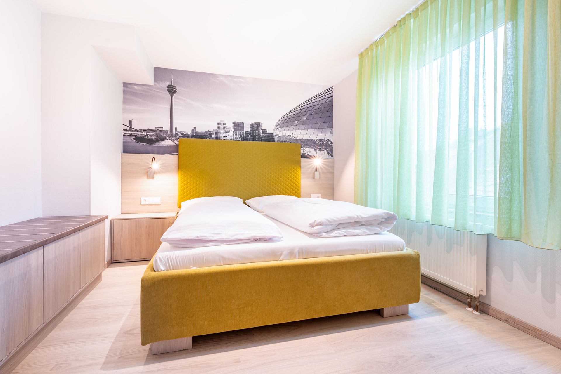 Gelbes Bett in einem modernen Zimmer mit Stadtpanorama-Wandbild und transparenten Vorhängen.