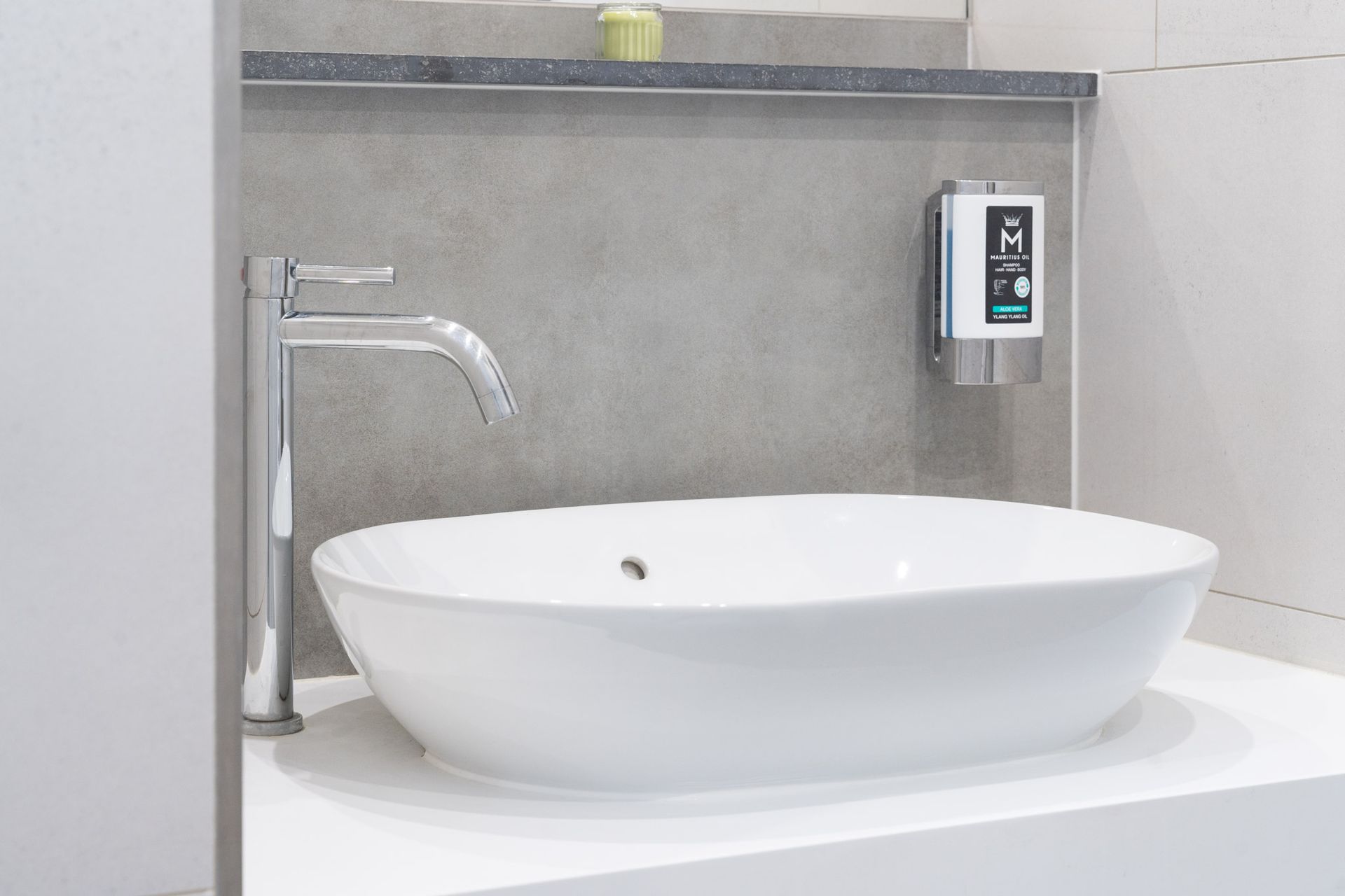 Waschbecken im Badezimmer mit verchromter Armatur, Seifenspender und weißem ovalem Becken vor einer grauen Wand.