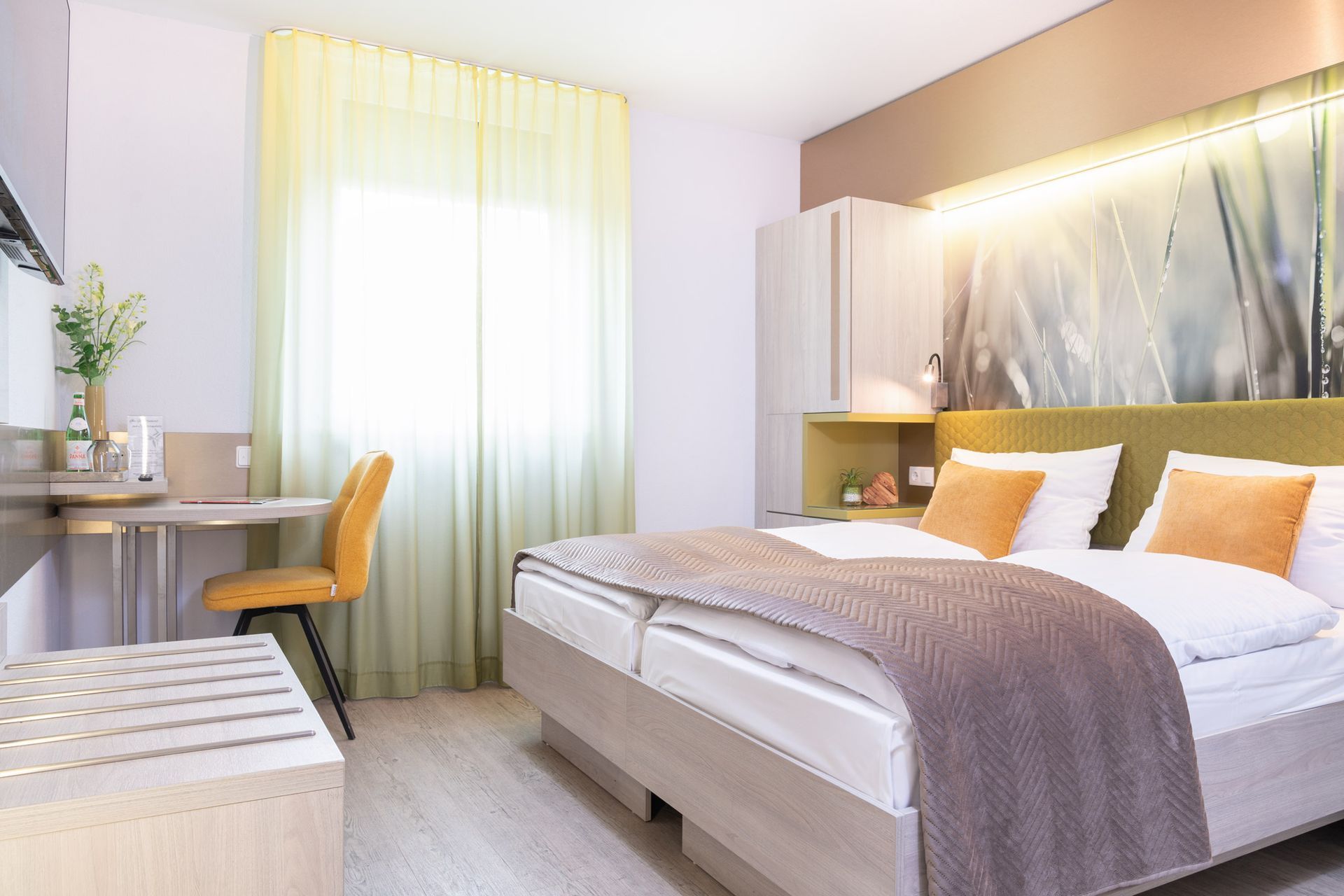 Hotelzimmer mit Bett, Schreibtisch und einem großen Fenster mit dünnen Vorhängen.