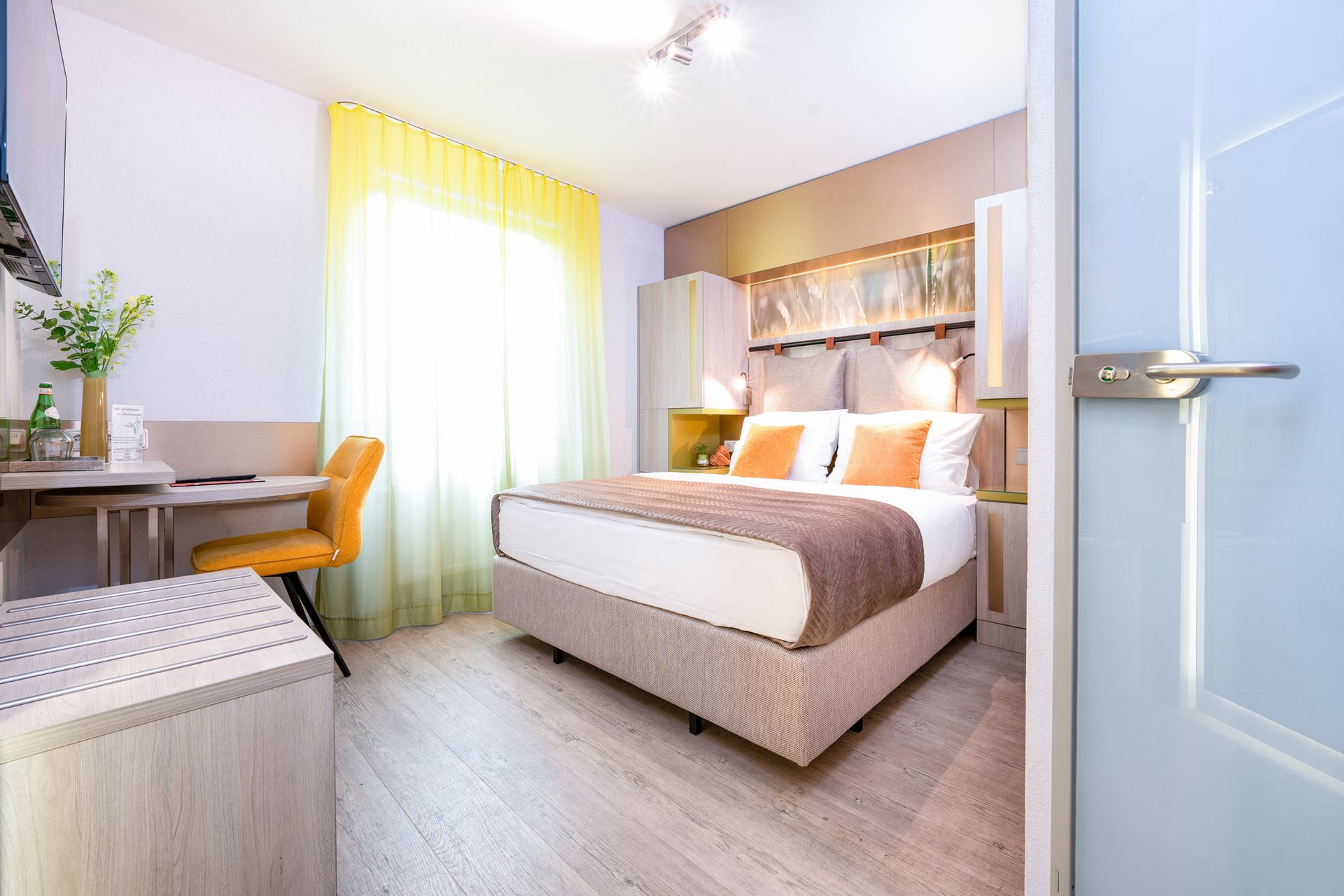 Schlafzimmer mit Bett, Schreibtisch und Fenster mit gelben Vorhängen. Helle Farbgestaltung.