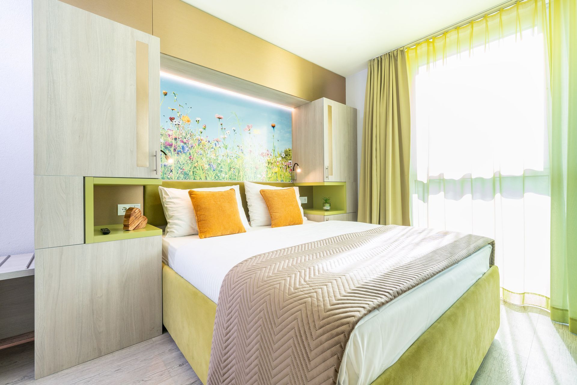 Schlafzimmer mit Bett, orangefarbenen Kissen, geblümtem Kopfteil und gelben Vorhängen.