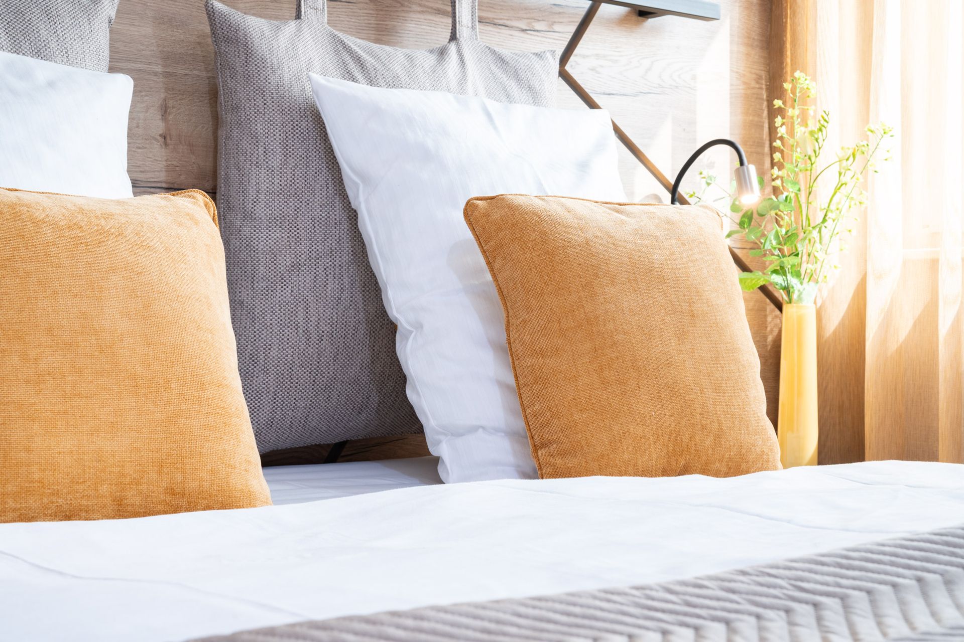 Bett mit Kissen in Weiß-, Grau- und Goldtönen. Eine Blumenvase steht daneben.