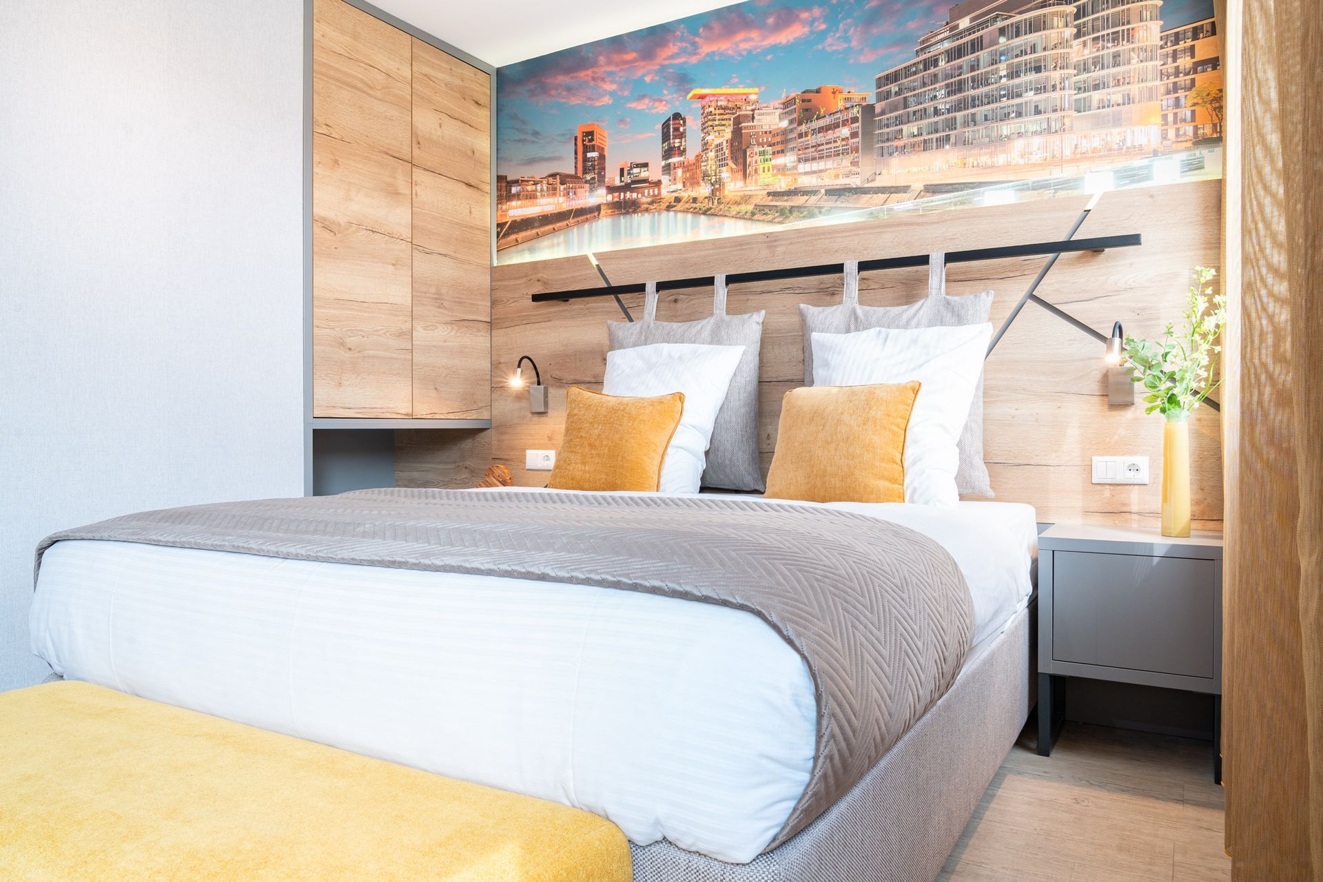 Hotelzimmer mit Bett, Bank, Holzwand und Stadtpanorama-Wandbild.