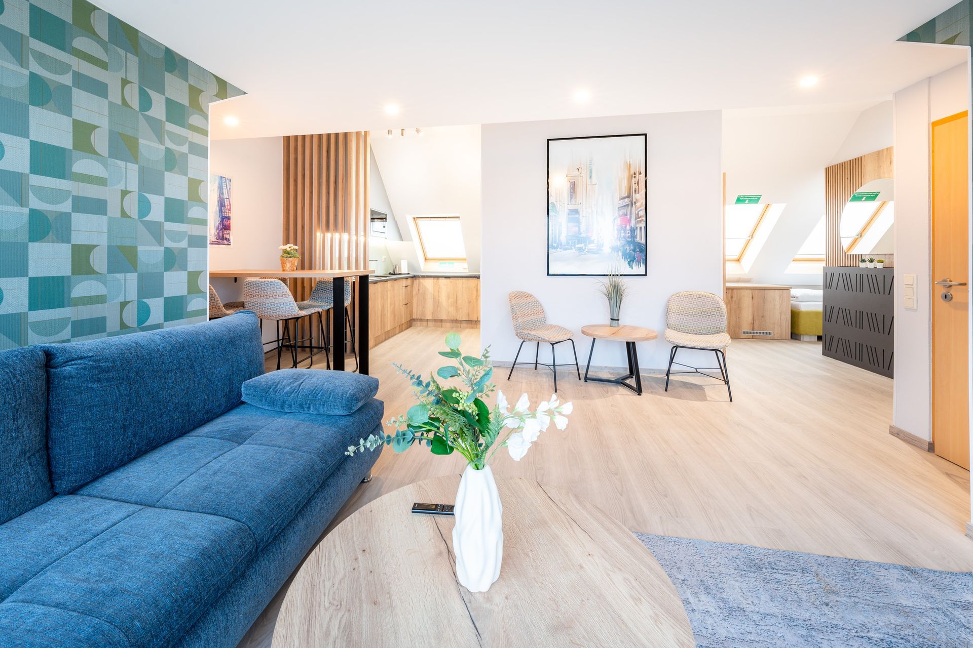 Heller, moderner Wohnraum. Blaues Sofa, Holzboden, Küche im Hintergrund. Helle Einrichtung mit Kunst.