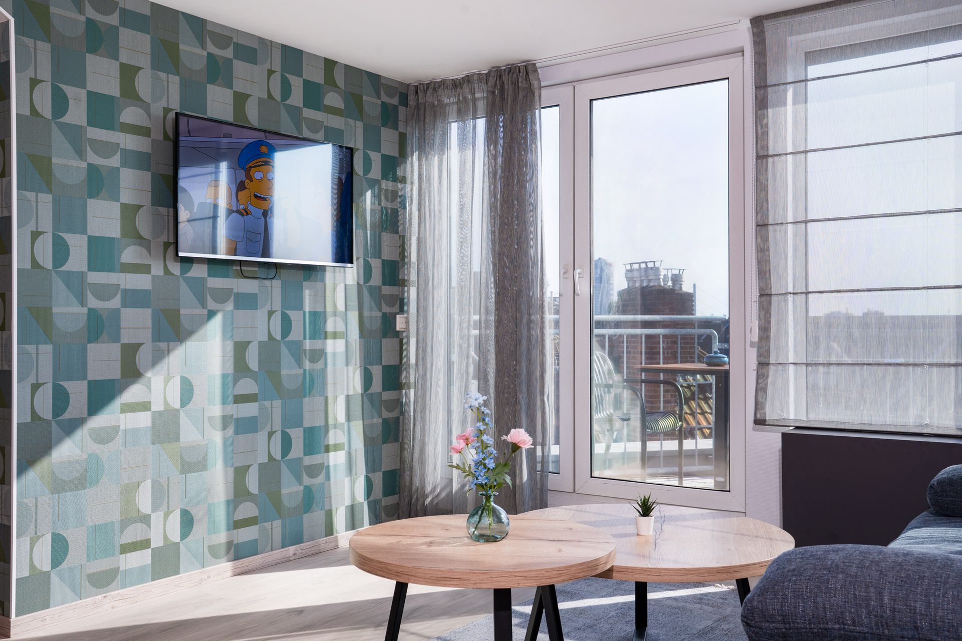 Wohnzimmer mit türkis gemusterter Wand, Fernseher, Couchtisch aus Holz und großem Fenster mit Balkonblick.