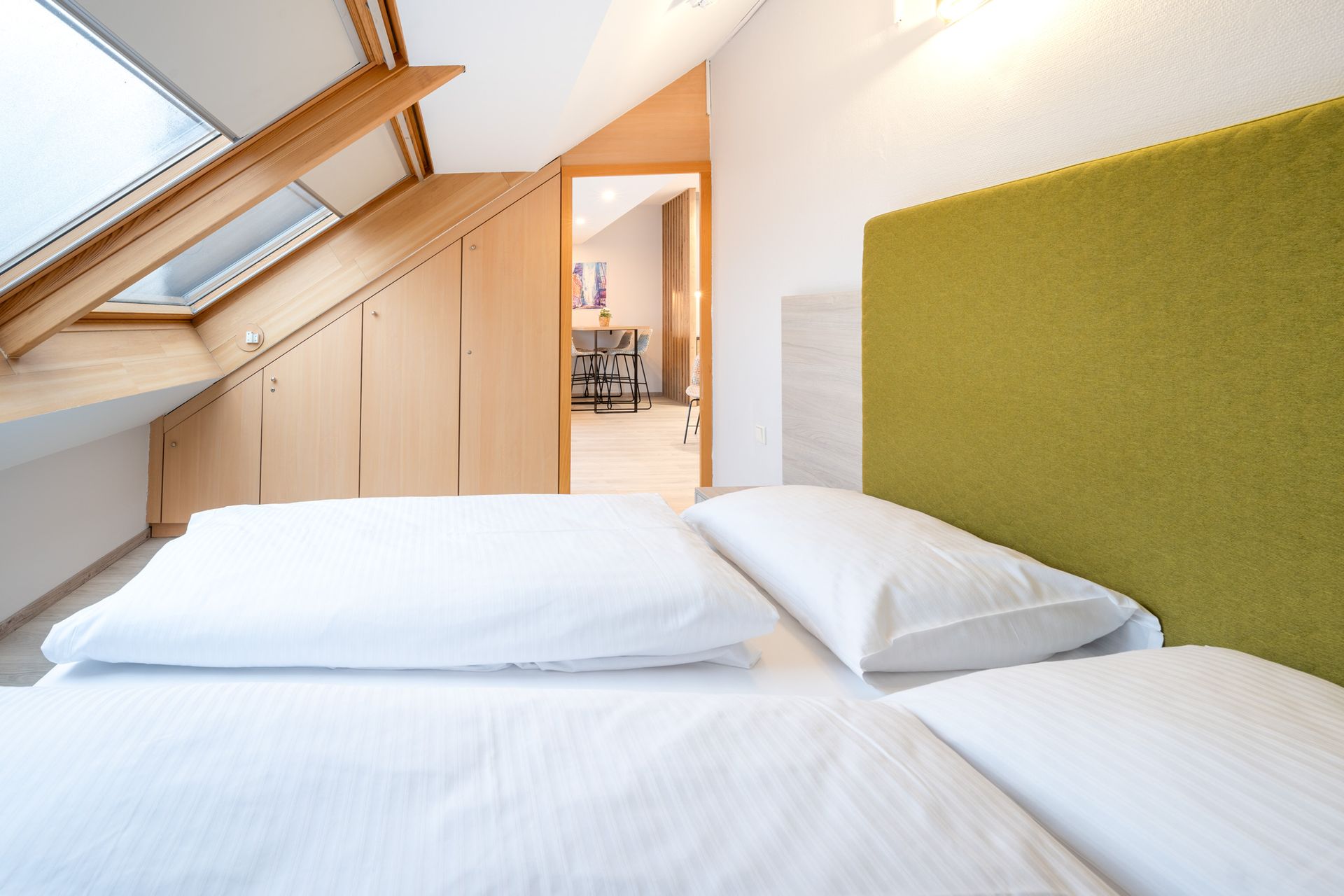 Schlafzimmer mit weißer Bettwäsche, grünem Kopfteil und Dachfenster. Tür zu einem weiteren Zimmer.