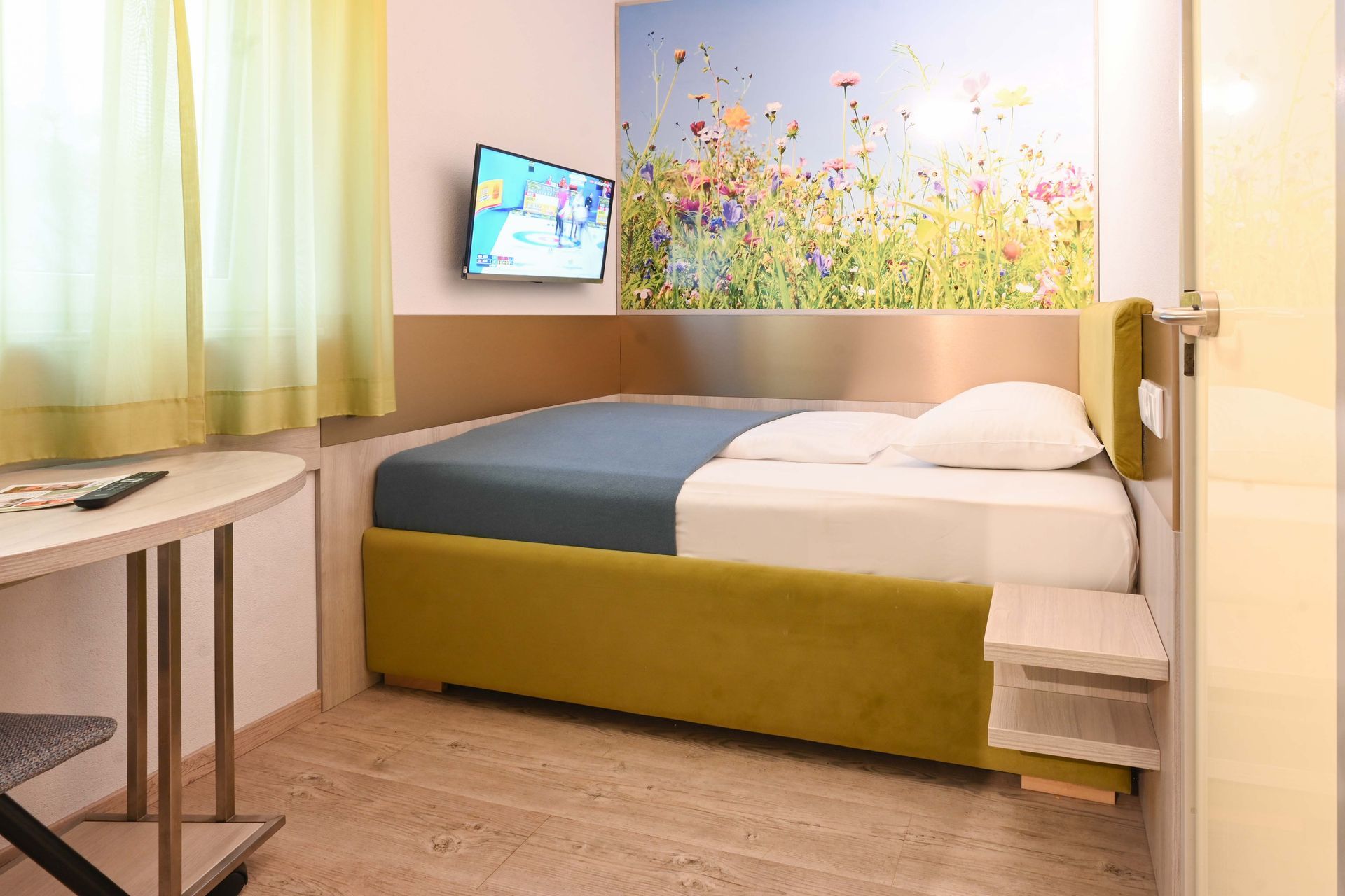 Kleines Hotelzimmer mit Bett, Fernseher, Schreibtisch und Blumenwandbild.