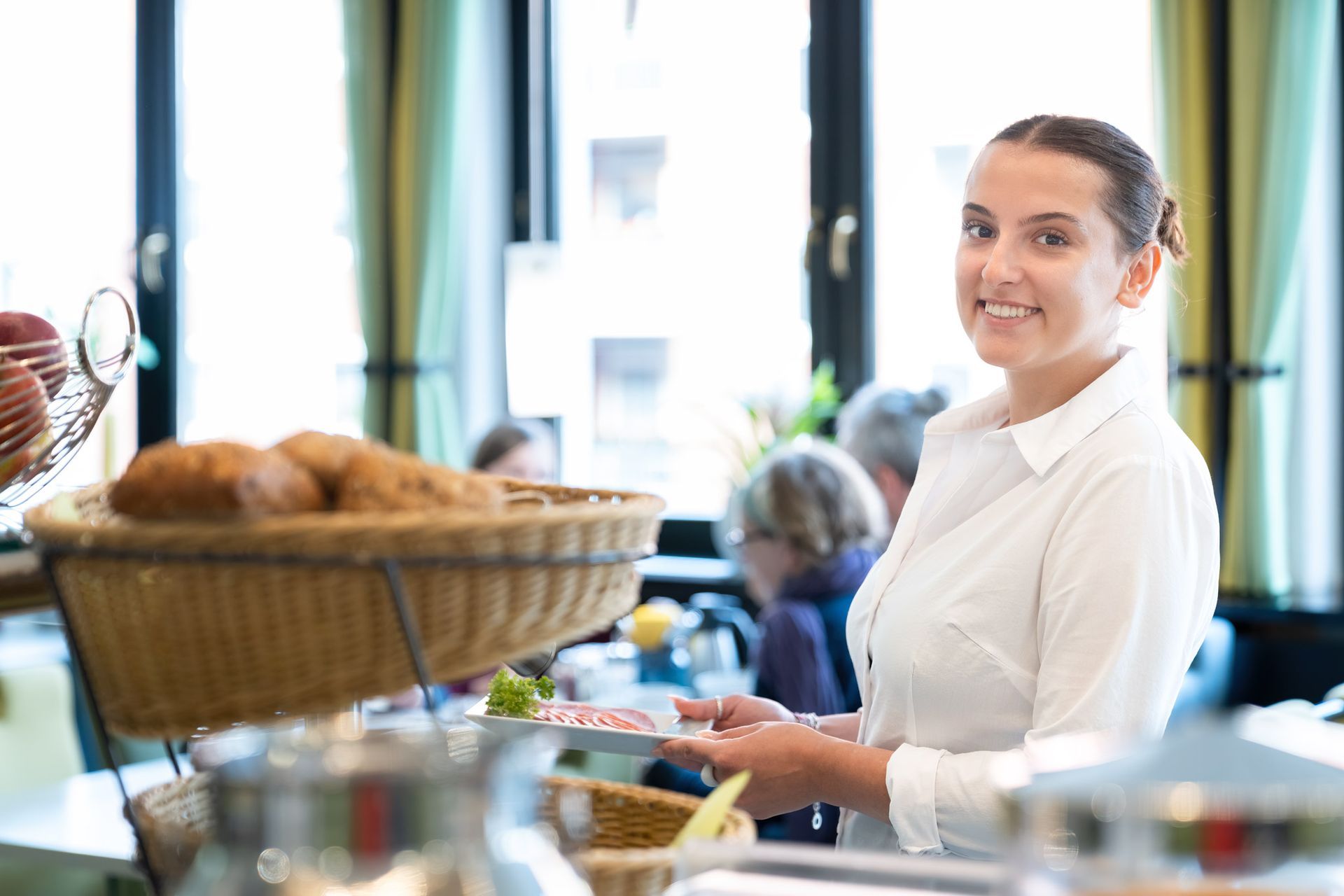 Eine Frau in weißem Hemd lächelt und hält einen Teller an einem Buffet. Im Hintergrund sind Brotkörbe und Menschen zu sehen.
