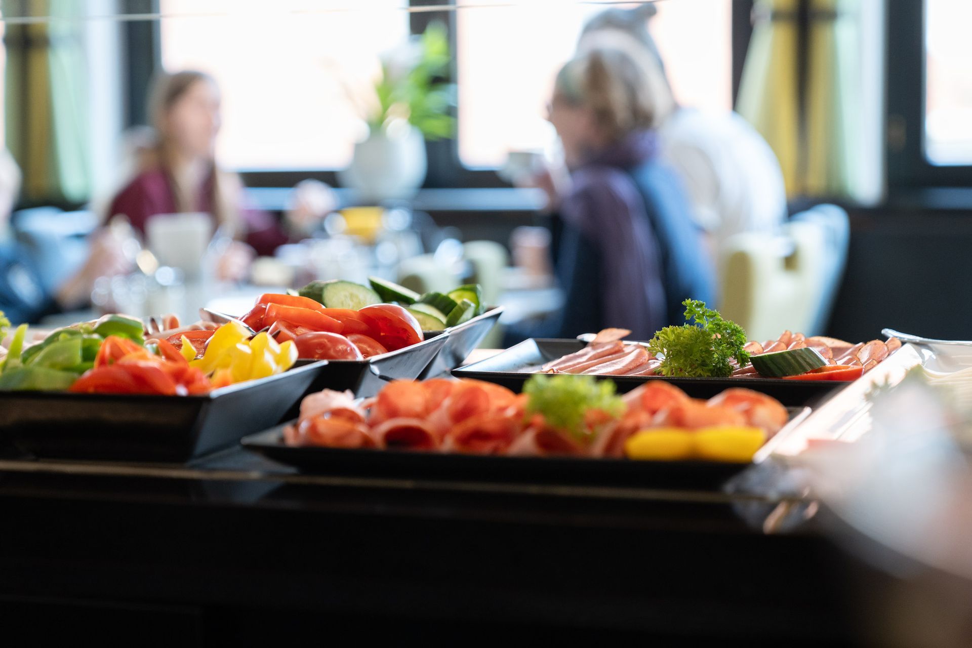 Buffet mit geschnittenem Gemüse, Fleisch und im Hintergrund Menschen, die an einem Tisch essen.