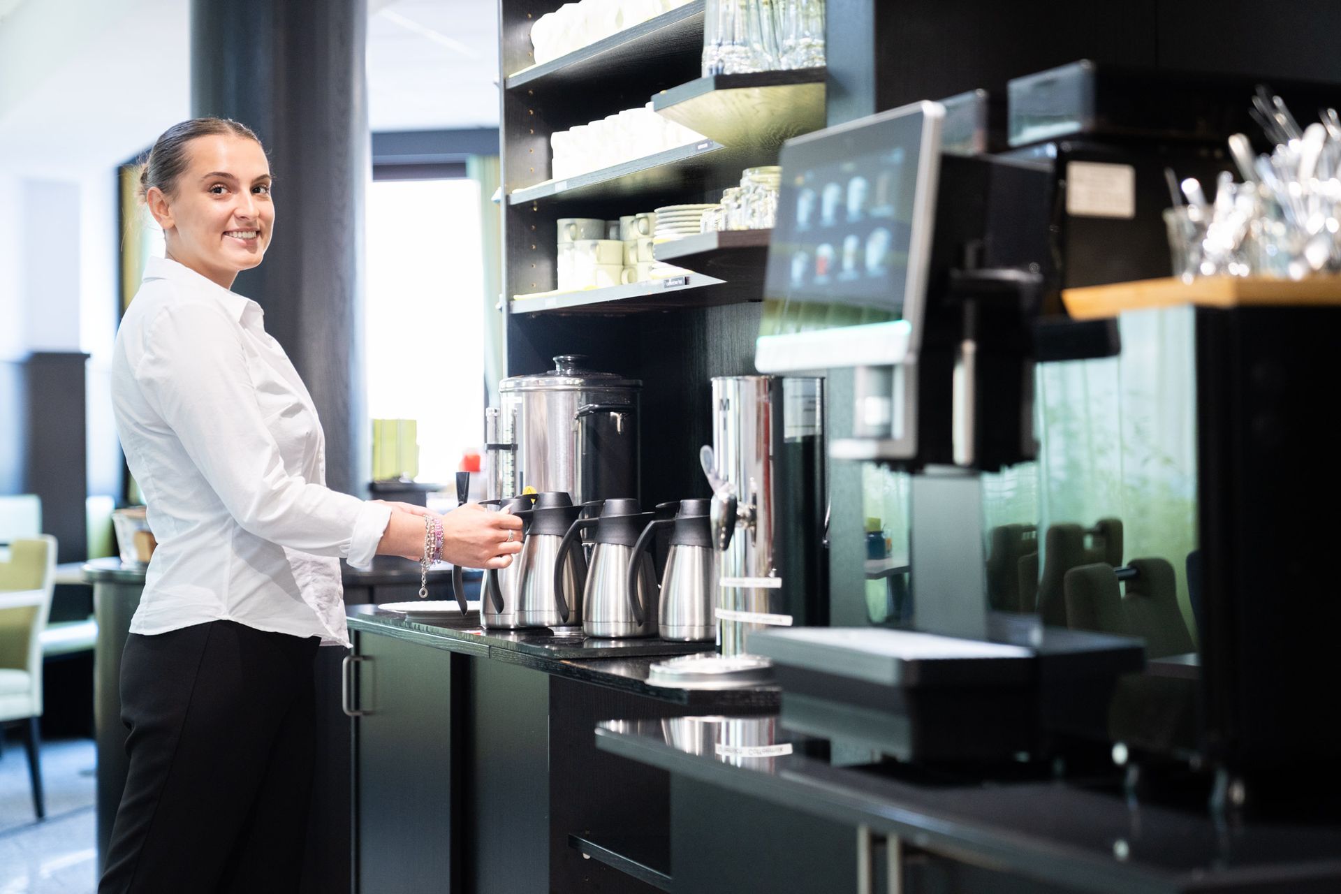 Frau in weißem Hemd an einer Kaffeestation, lächelt und bereitet Kaffee zu.