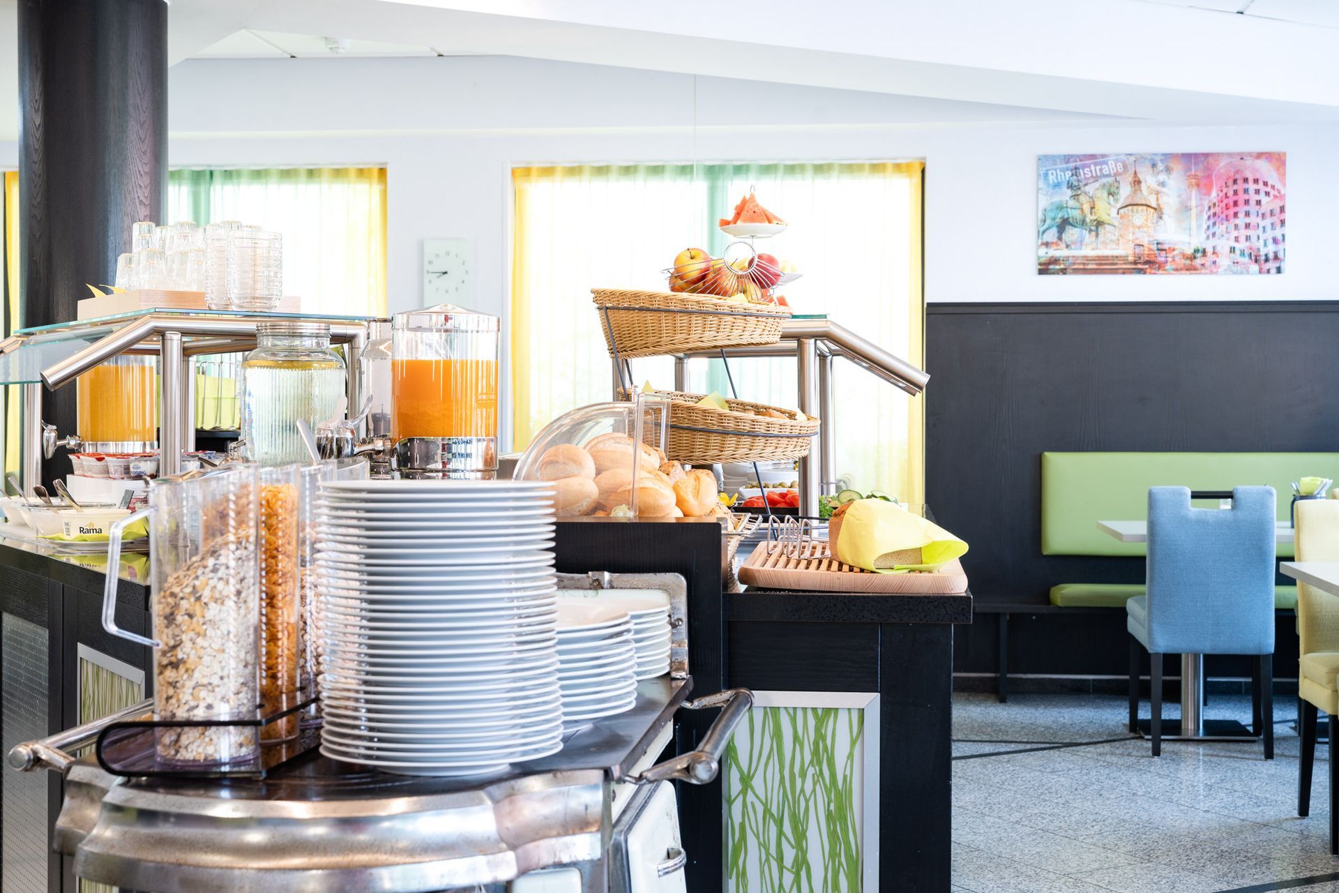 Frühstücksbuffet mit Saft, Müsli, Obst, Eiern und Tellern. Essbereich mit Tischen und Stühlen.