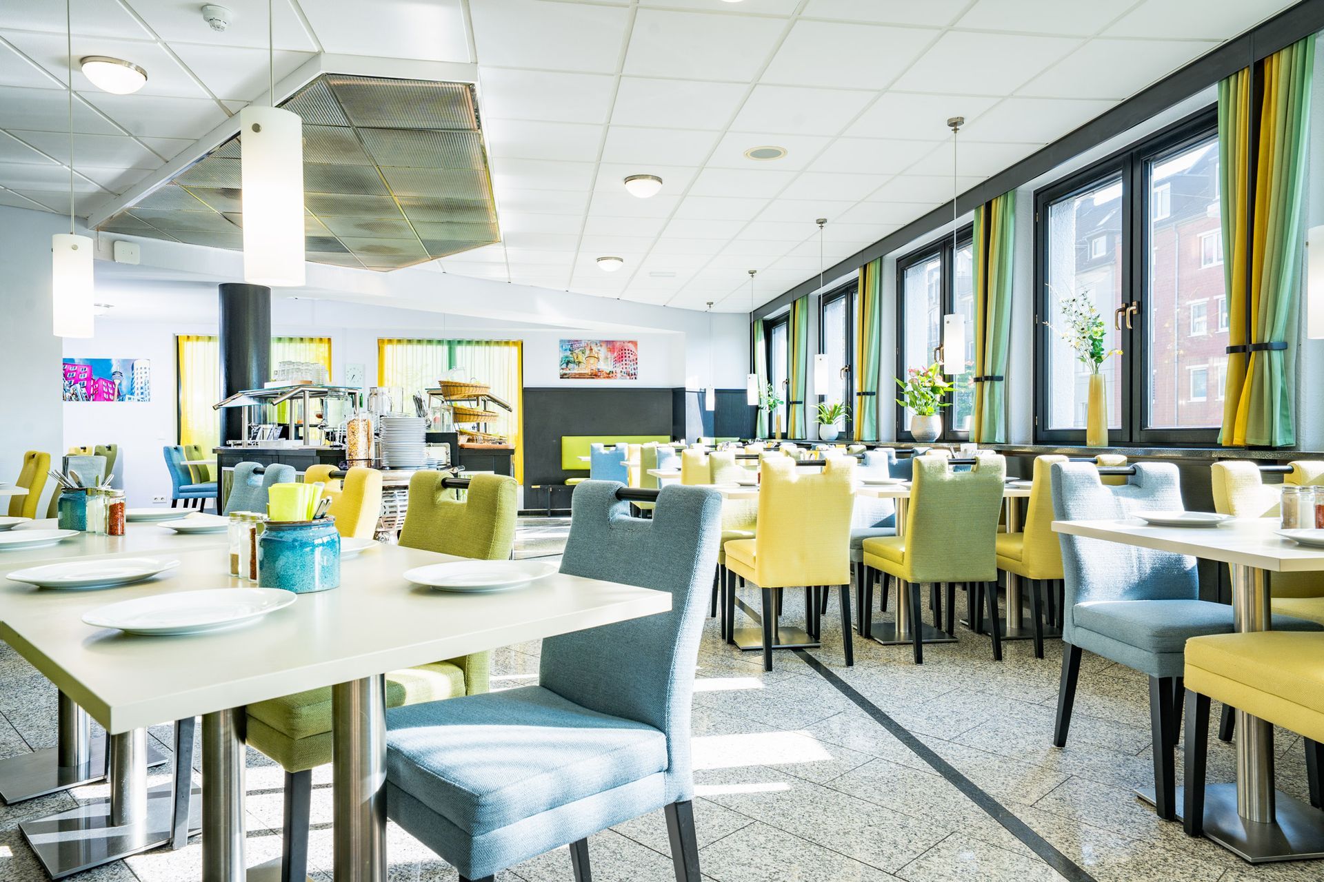 Esszimmer mit Tischen und farbenfrohen Stühlen, großen Fenstern mit gelben und blauen Vorhängen und Buffetbereich.