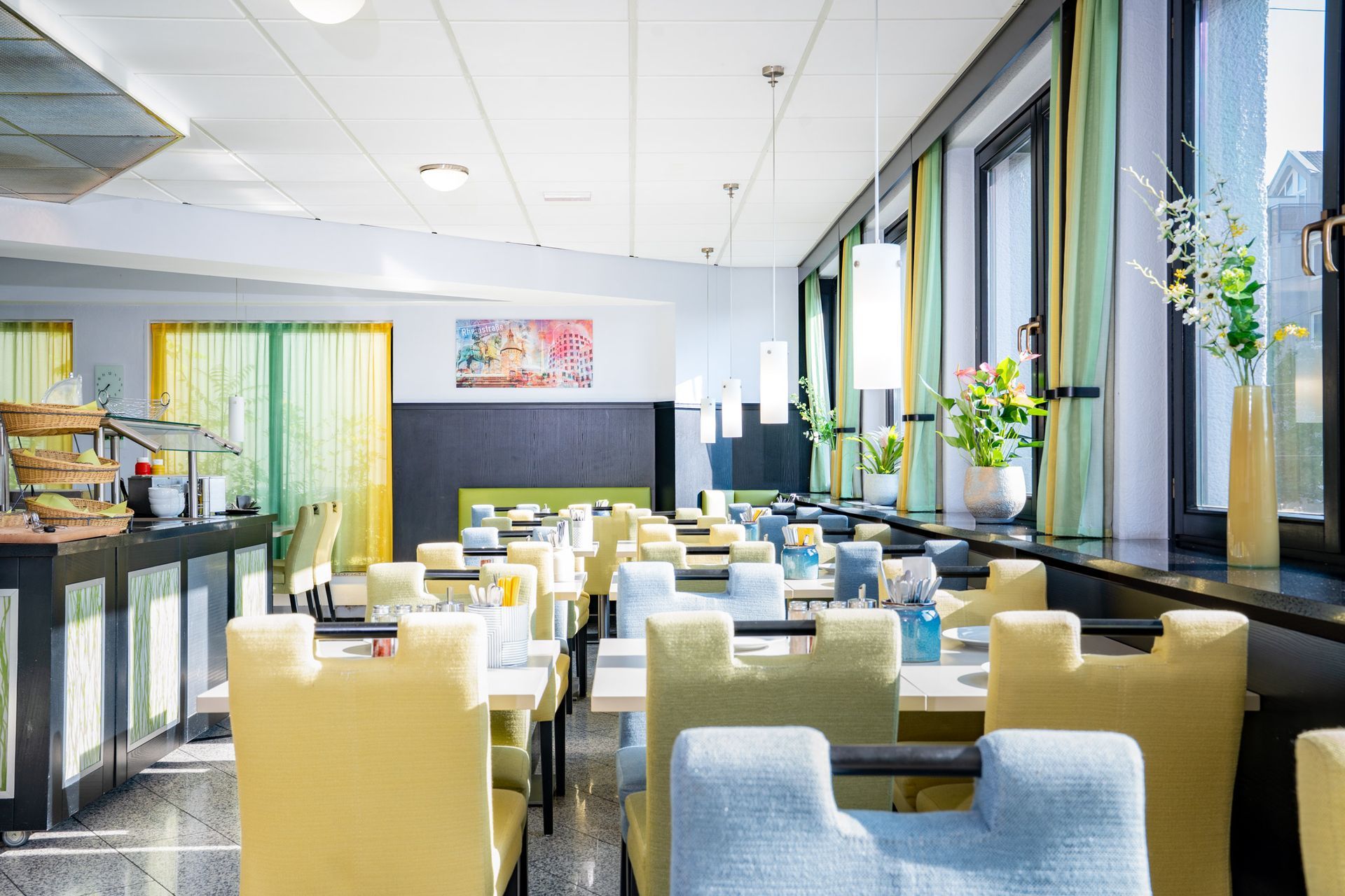 Restaurant-Essbereich mit gedeckten Tischen. Gelbe und blaue Stühle säumen die Tische in Fensternähe.