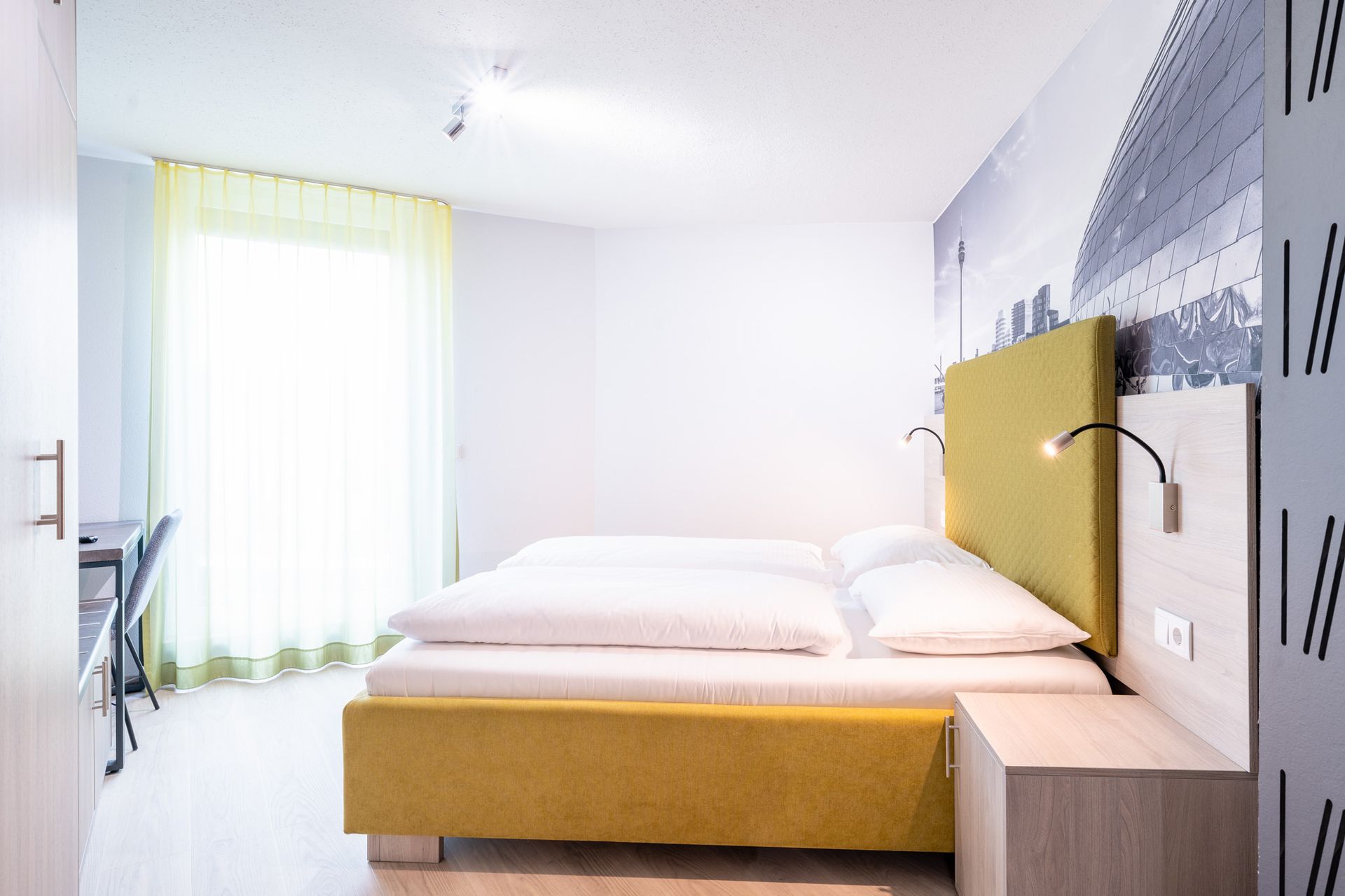 Schlafzimmer mit gelbem Bett, weißen Wänden, großem Fenster und Nachttisch.