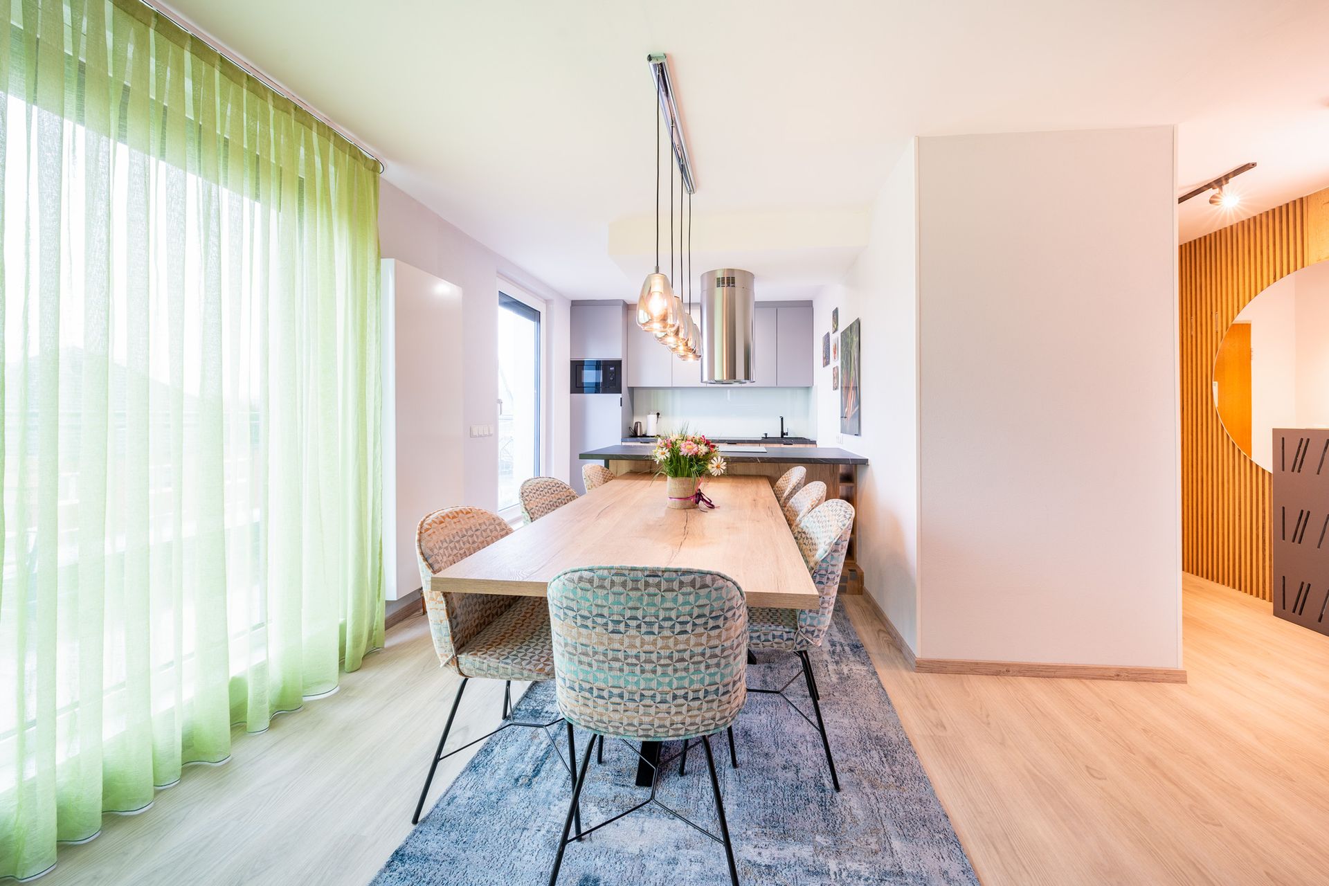 Essbereich mit langem Holztisch, Stühlen und Küche im Hintergrund. Grüne Vorhänge und helle Holzböden.