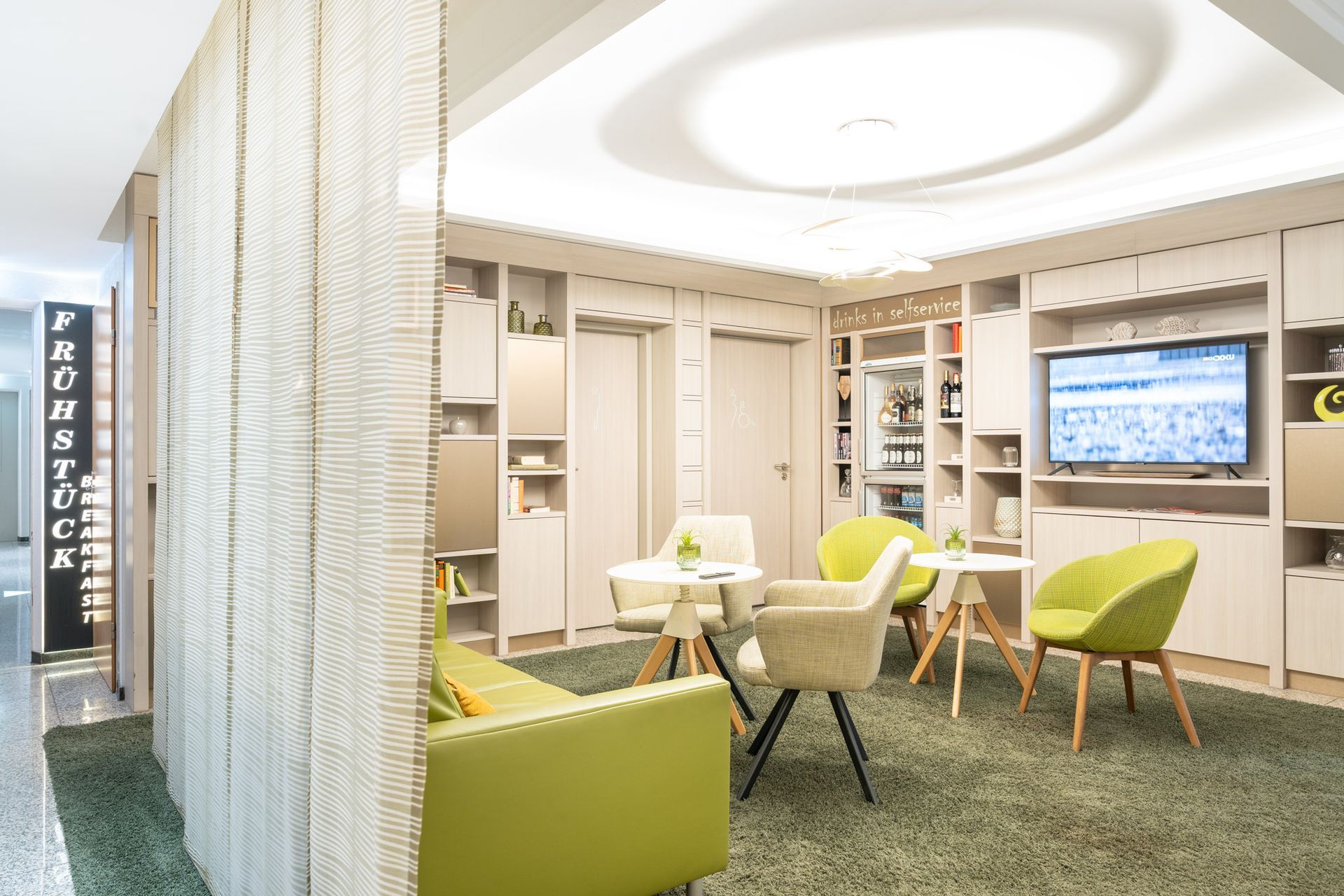 Moderner Loungebereich mit grünen und neutralen Möbeln, Fernseher und dekorativer Raumteilerwand.