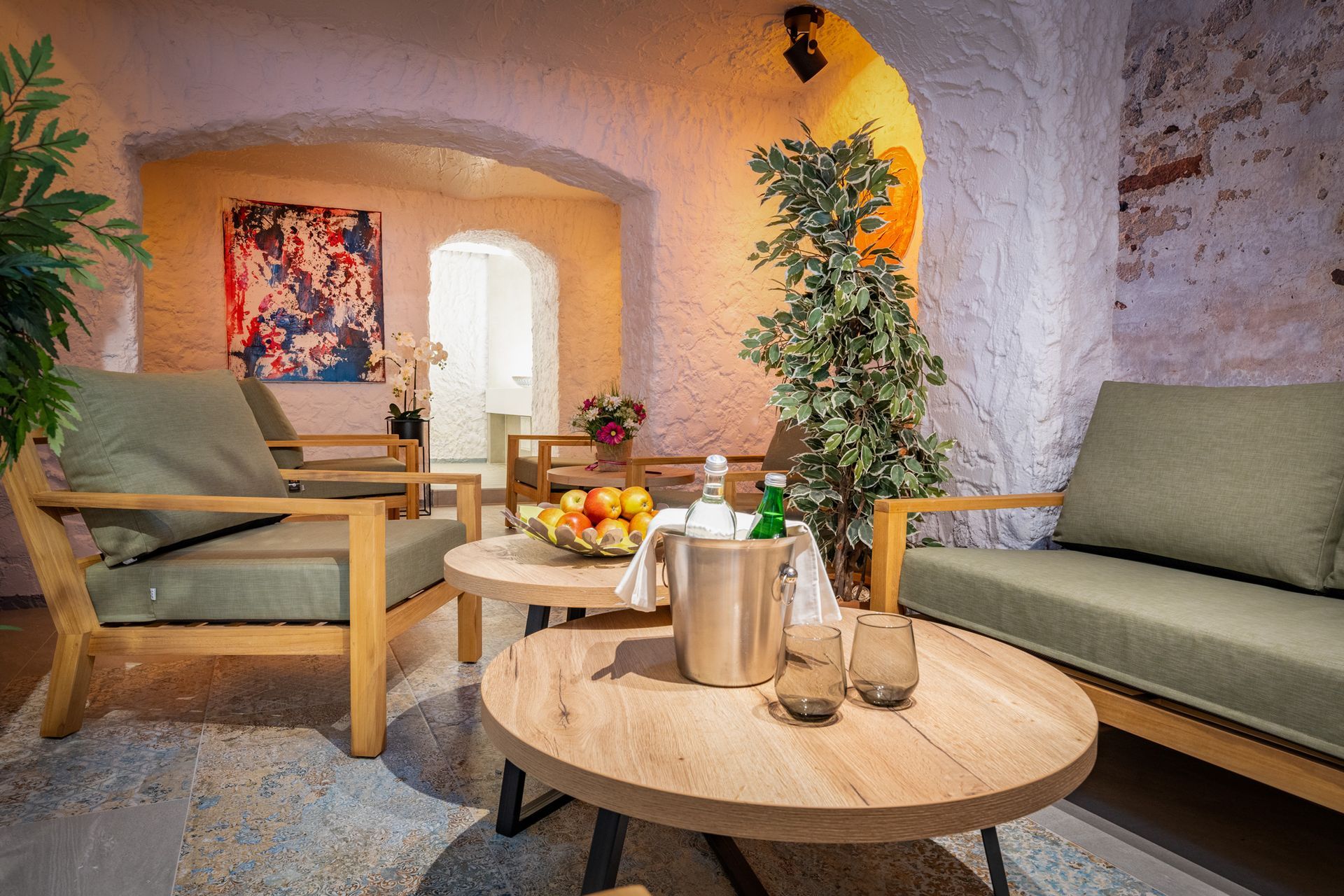 Gemütliche Sitzecke mit Holztischen, olivgrünen Kissen und einem Champagnerkühler. Weiß getünchte Wände, natürliches Licht.