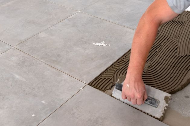 Une personne utilise une truelle crantée pour étaler une fine couche de mortier colle sur un sol avant la pose de grands carreaux.