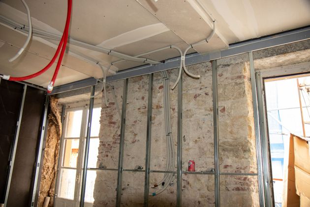 Intérieur d'un chantier de construction avec une structure métallique contre un mur en pierre, des câbles apparents et un plafond fraîchement enduit.