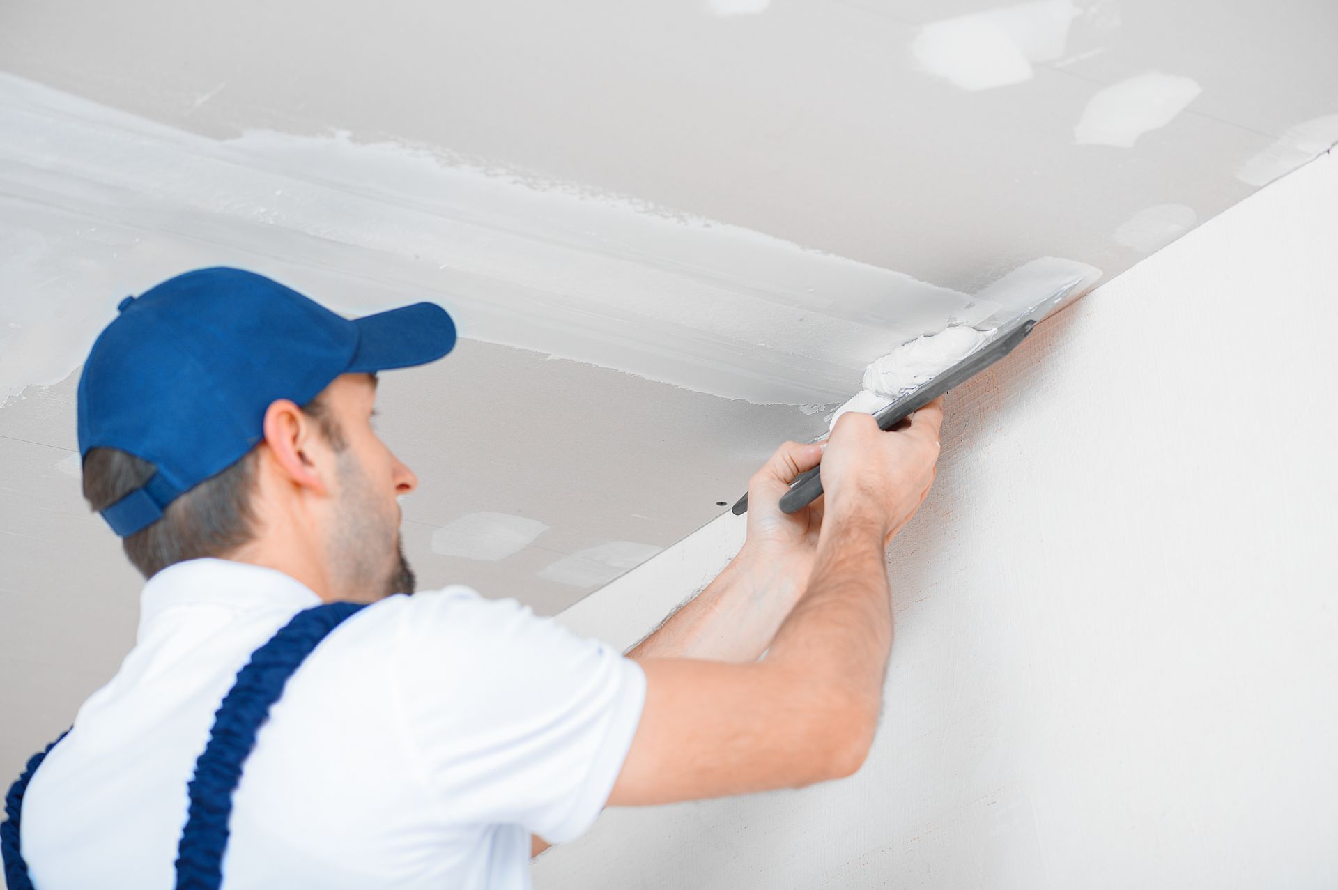 Un professionnel, coiffé d'une casquette et vêtu d'une salopette de travail, applique un enduit à joints sur un angle de plafond à l'aide d'un couteau à mastic métallique.
