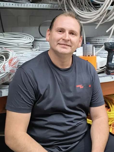 Ein Mann sitzt in einer Werkstatt neben aufgewickelten Kabeln und Geräten; er trägt ein dunkles T-Shirt und lächelt leicht.