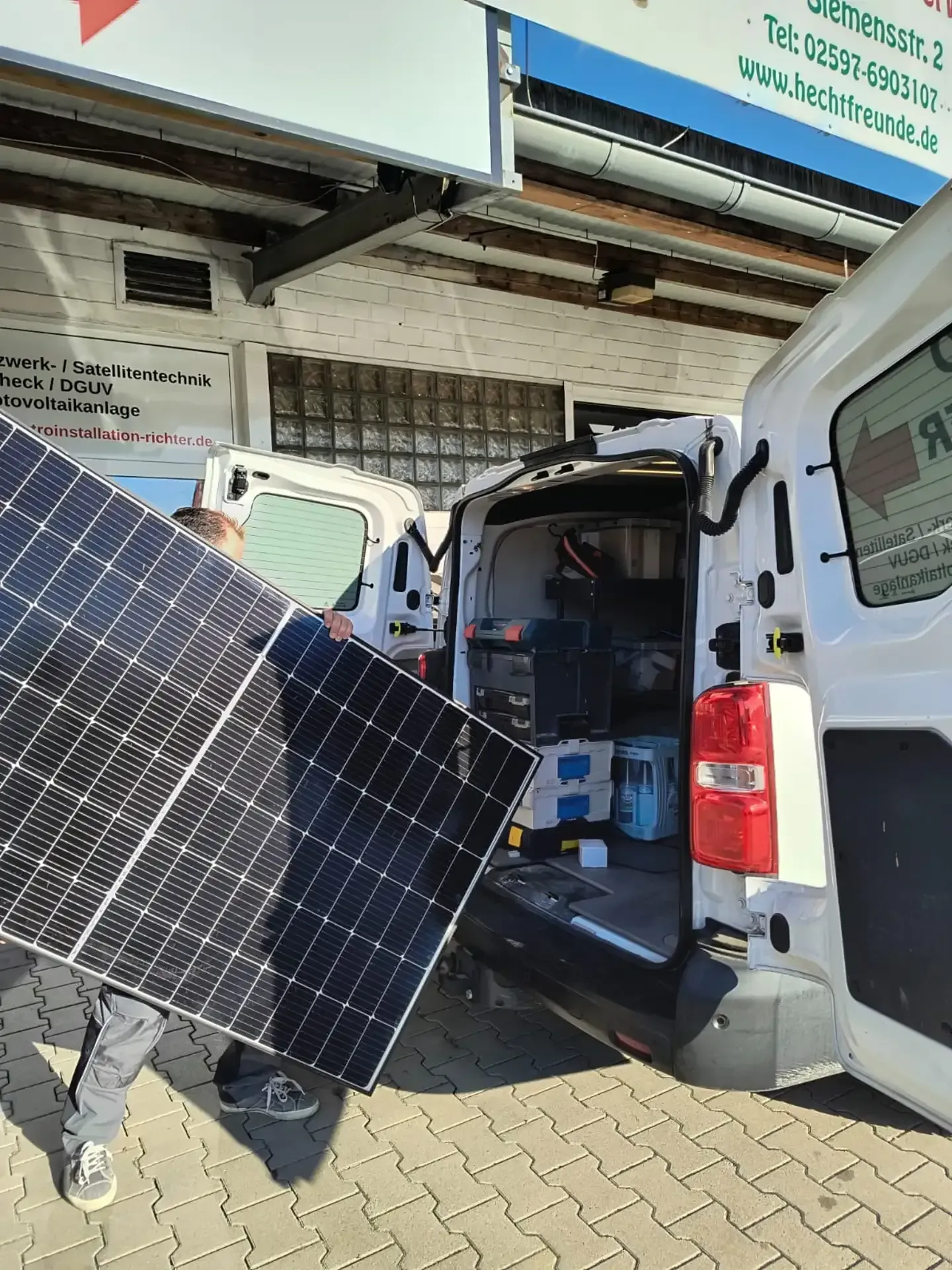 Eine Person trägt ein Solarpanel neben einem weißen Lieferwagen mit geöffneten Hecktüren vor einem Gebäude.