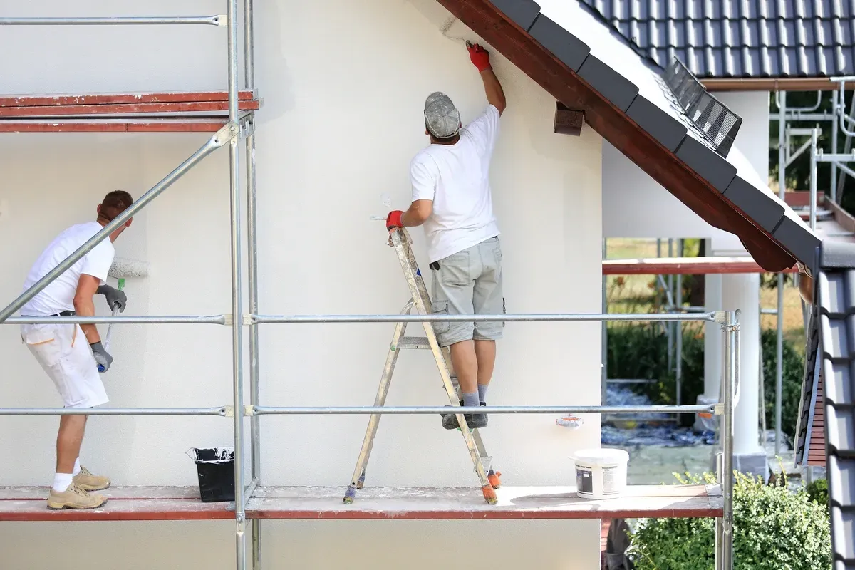 Zwei Männer streichen auf einem Gerüst die Seite eines Hauses.