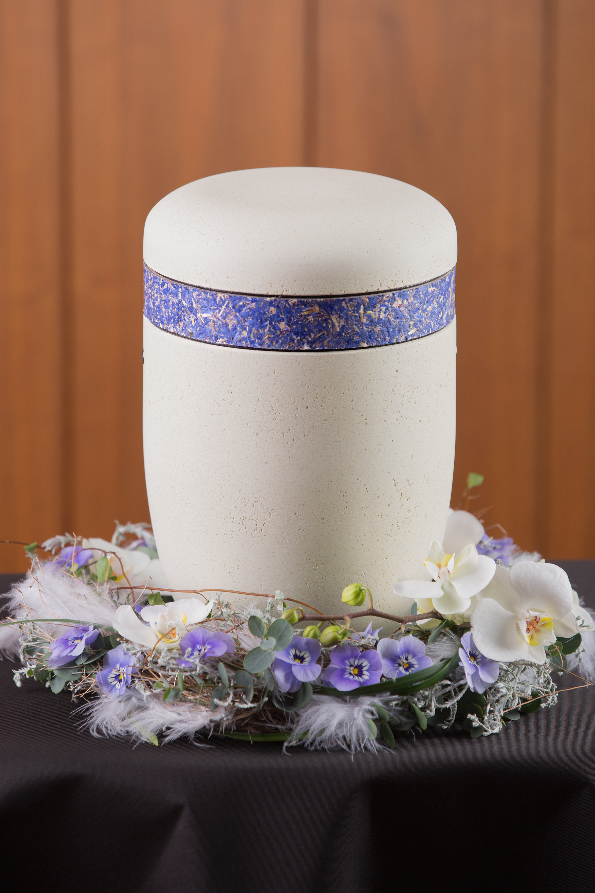 Helle Urne mit blauem Rand, darunter ein besonderes Gesteck mit Federn, Eukalyptus, violetten Pfingst-Veilchen und weißen Orchideen