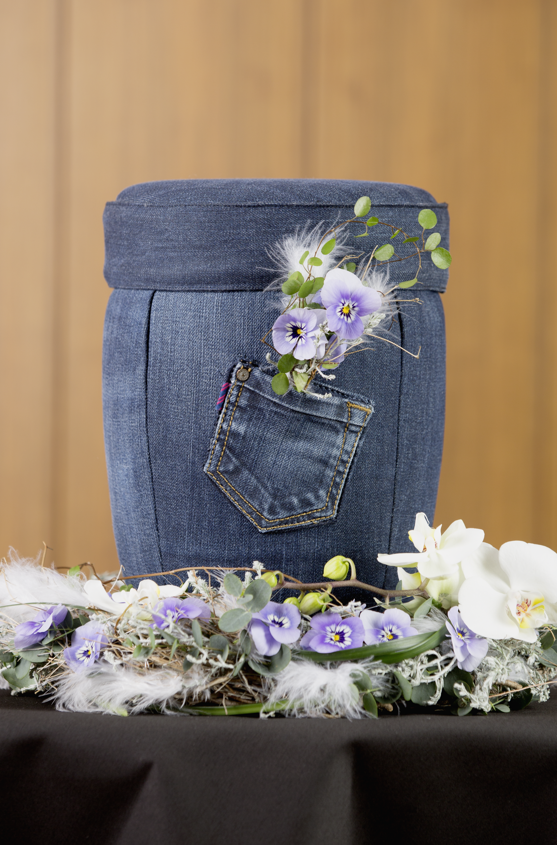Außergewöhnliche Jeans Urne, darunter ein besonderes Gesteck mit Federn, Eukalyptus, violetten Pfingst-Veilchen und weißen Orchideen