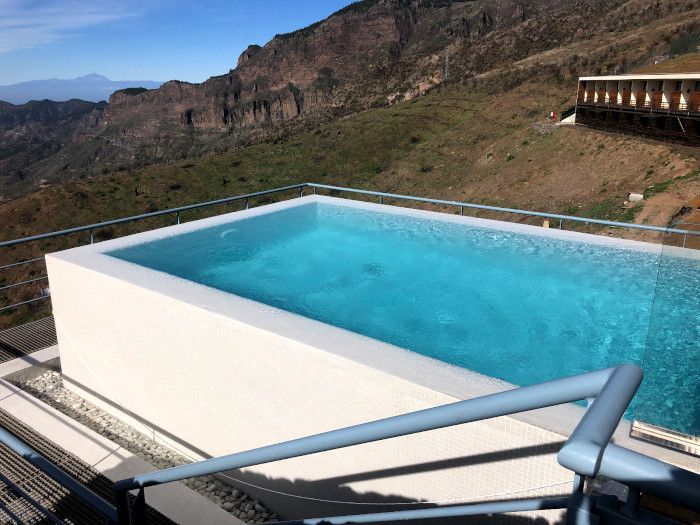 Una gran piscina está situada en la cima de una colina con montañas al fondo.